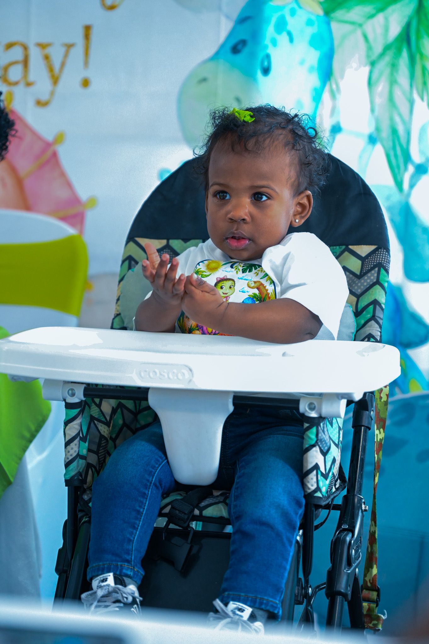 Cute baby boy clapping hands while seated in high chair at dinosaur-themed party.