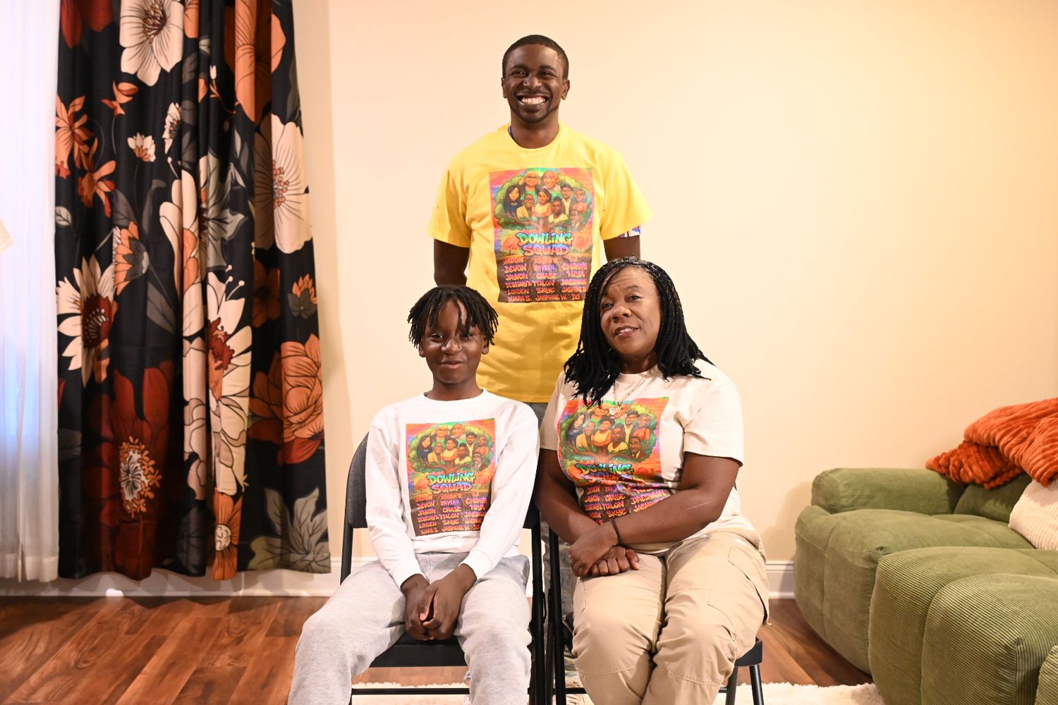 Three people wearing matching yellow graphic shirts pose together indoors, one seated in a wheelchair, in a cozy living room.