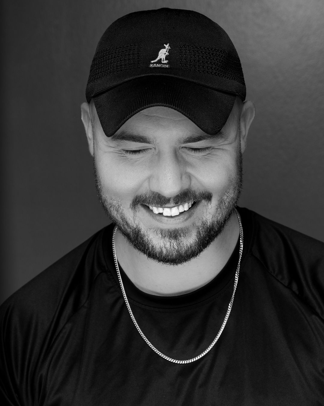 Black and white portrait of a smiling man with beard wearing a black Kangol cap and chain necklace.