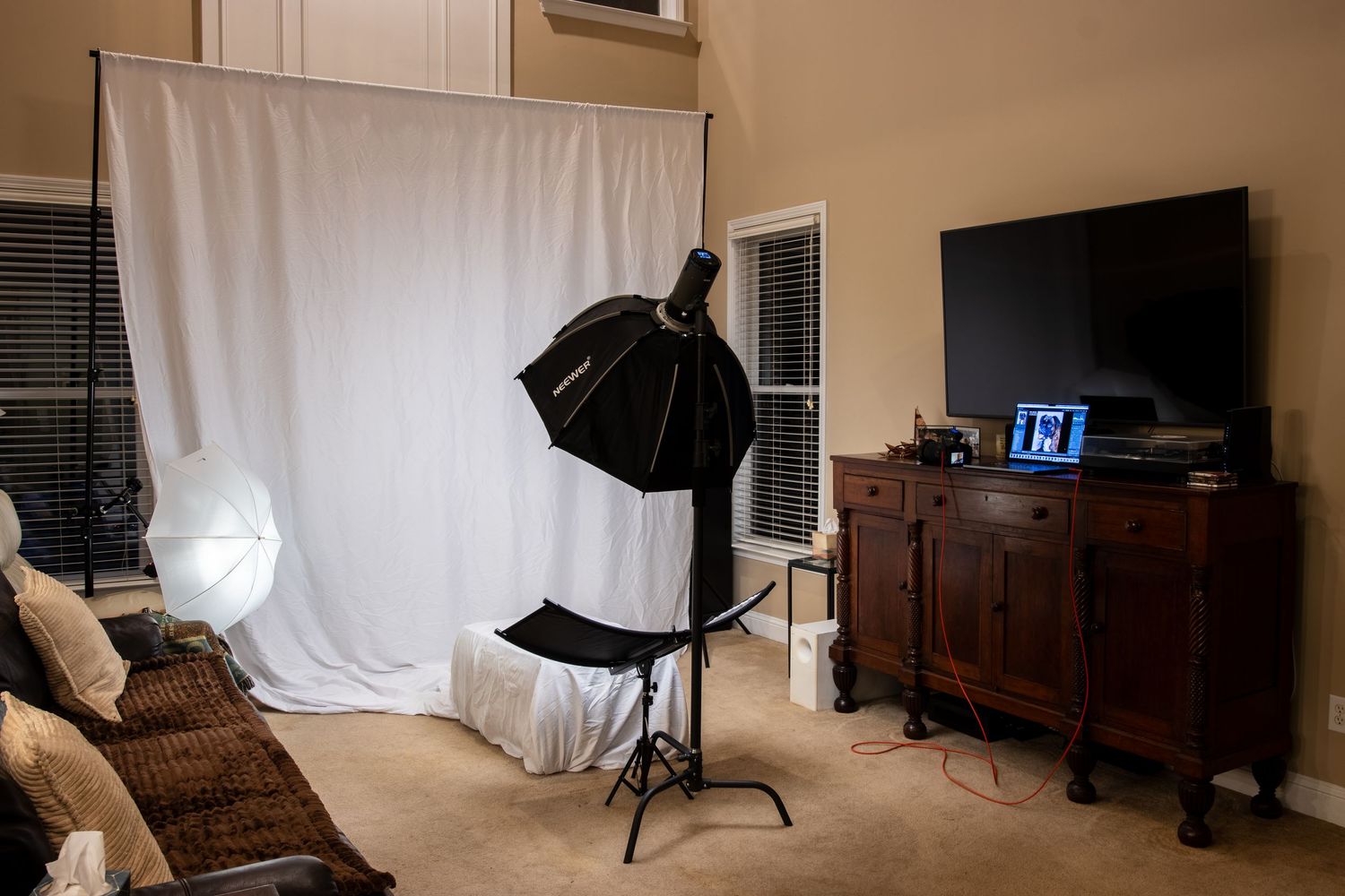 Home photography studio setup with white backdrop, softbox lighting, and a dark wood TV console in a living room.