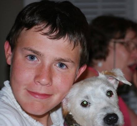 A smiling blue-eyed boy poses cheek-to-cheek with a white Jack Russell Terrier mix dog indoors.