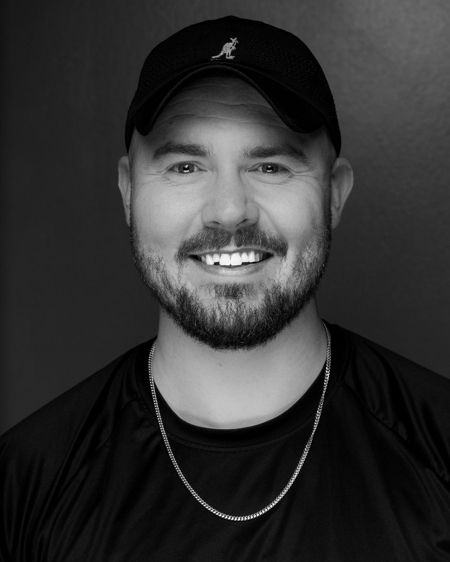 Black and white close-up portrait of a smiling bearded man in a black Kangol cap and chain necklace.