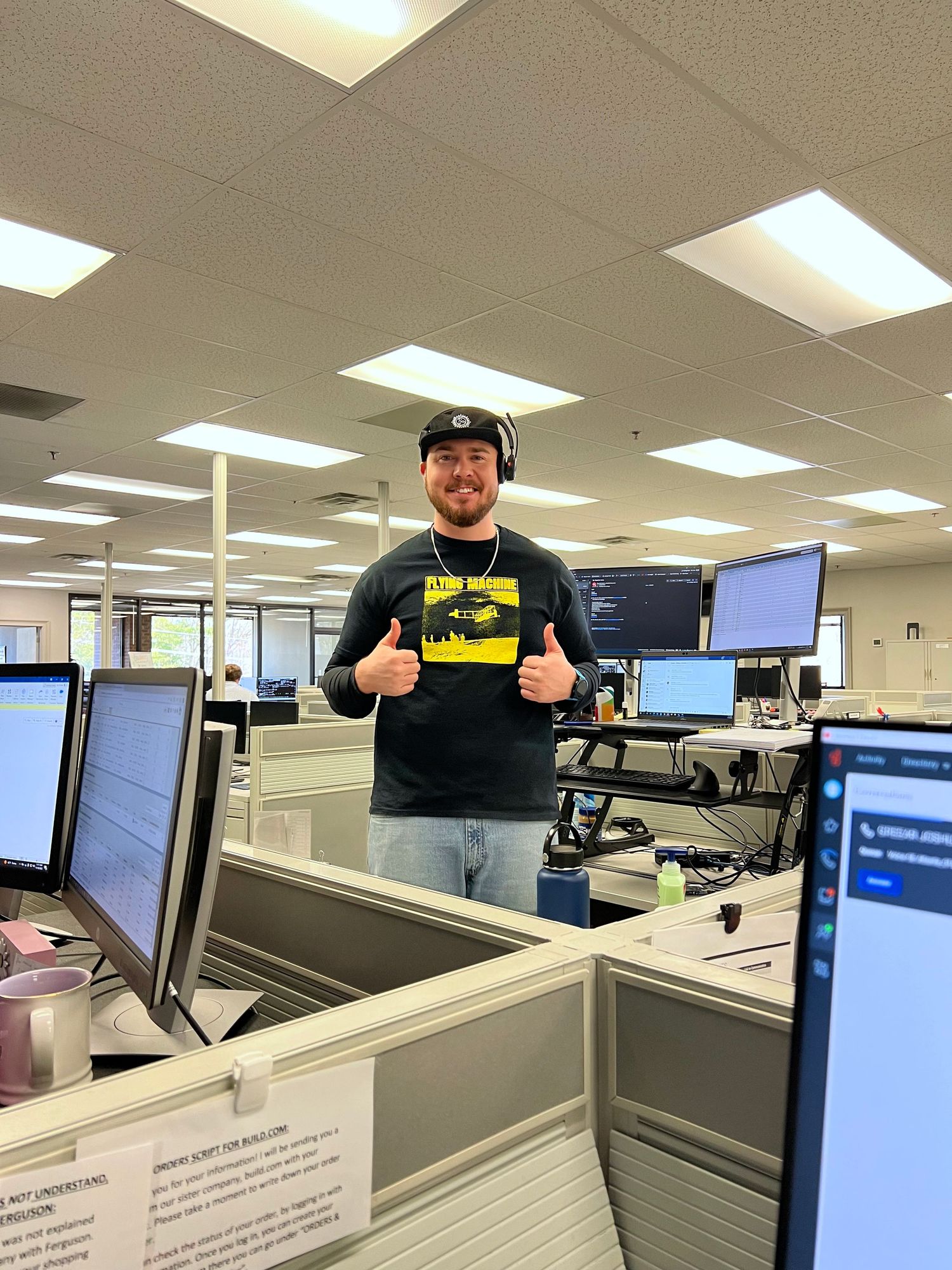Smiling man with headset on gives thumbs up in a modern office cubicle environment.