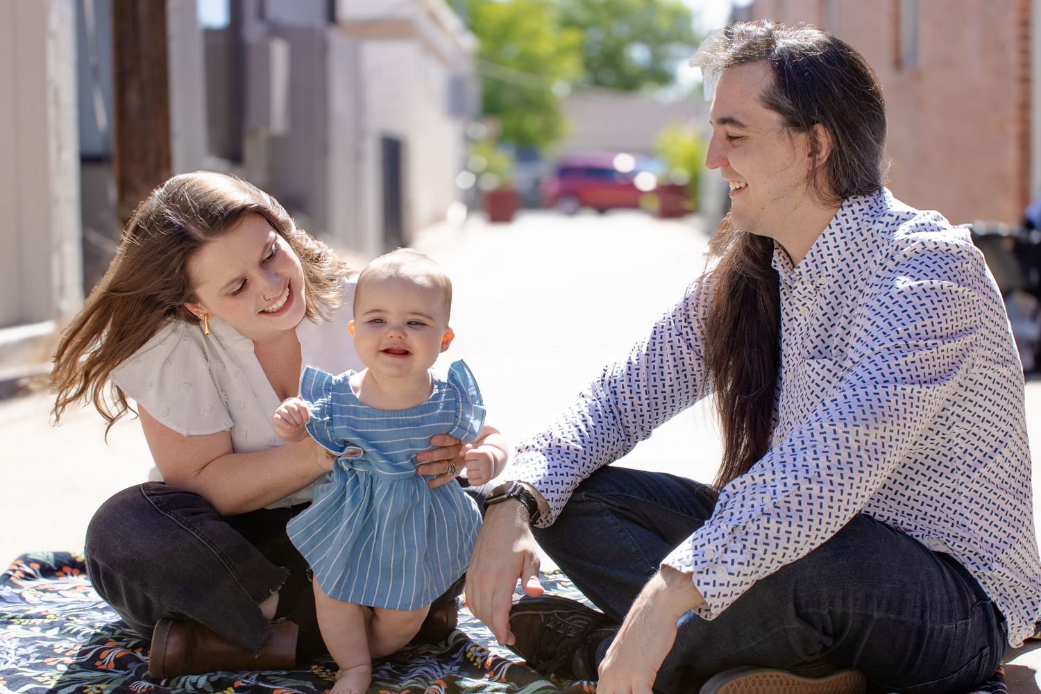 A couple sits outdoors on a blanket smiling at a happy baby girl in a blue dress.