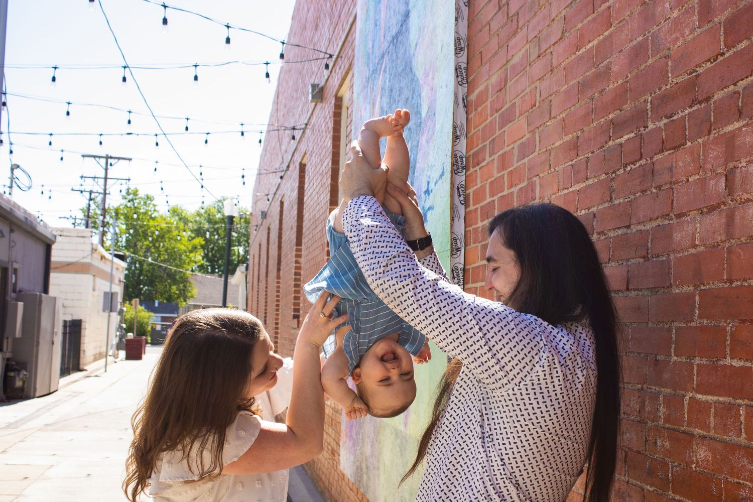 A couple playfully lift a baby against a colorful brick wall mural in a sunny urban alley.