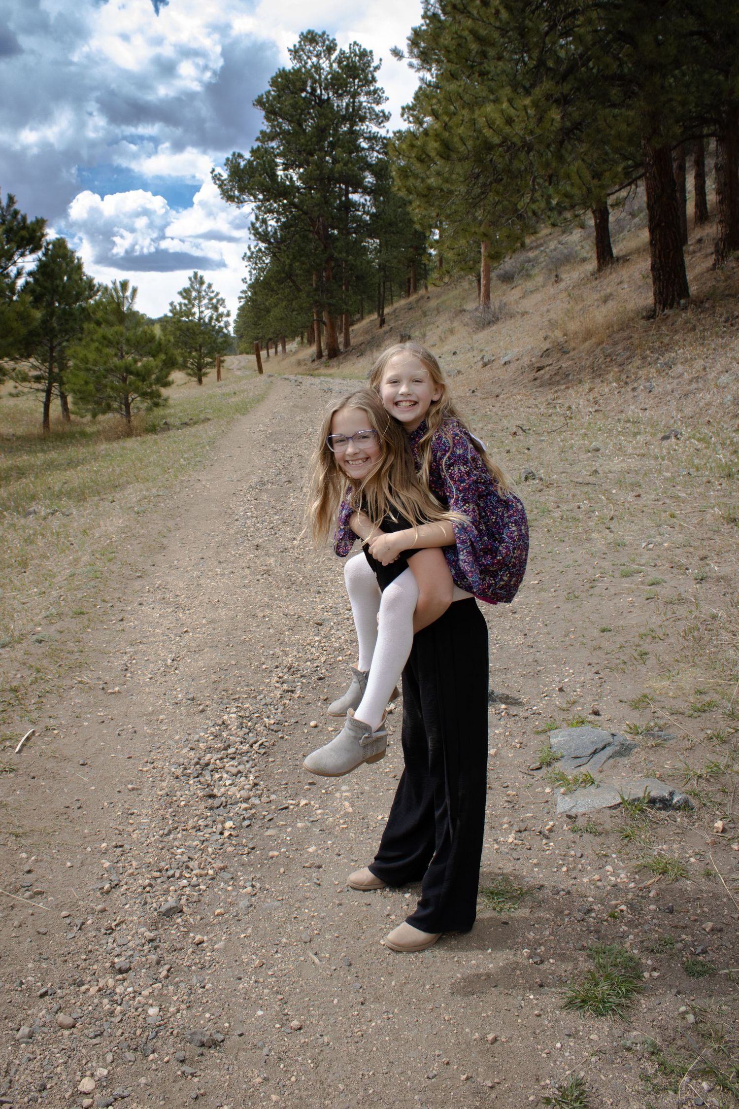 Two girls laugh as one gives the other a piggyback ride on a scenic mountain dirt trail surrounded by pine trees.