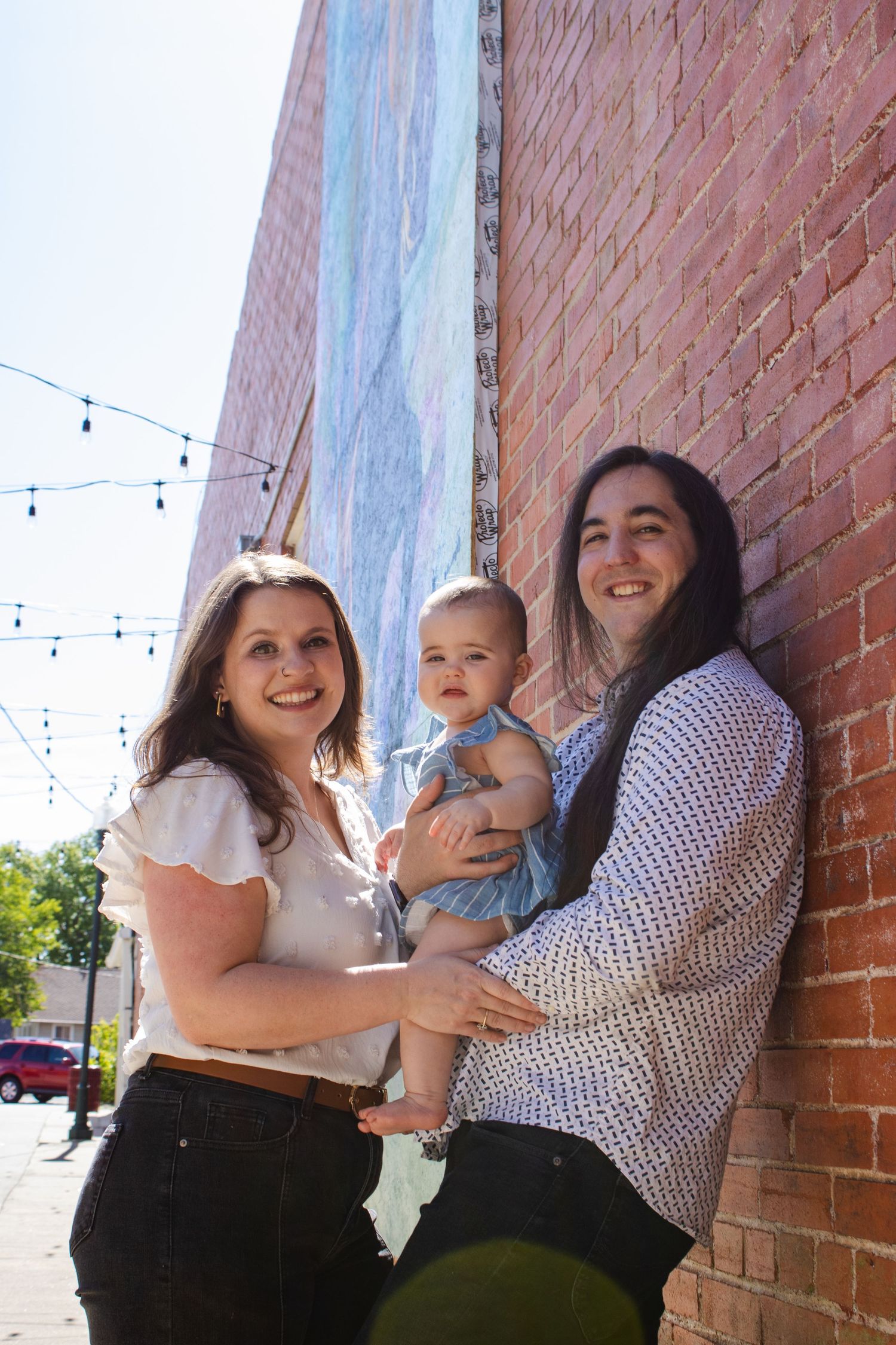 A couple hold a baby in a blue dress against a brick wall with a colorful mural.
