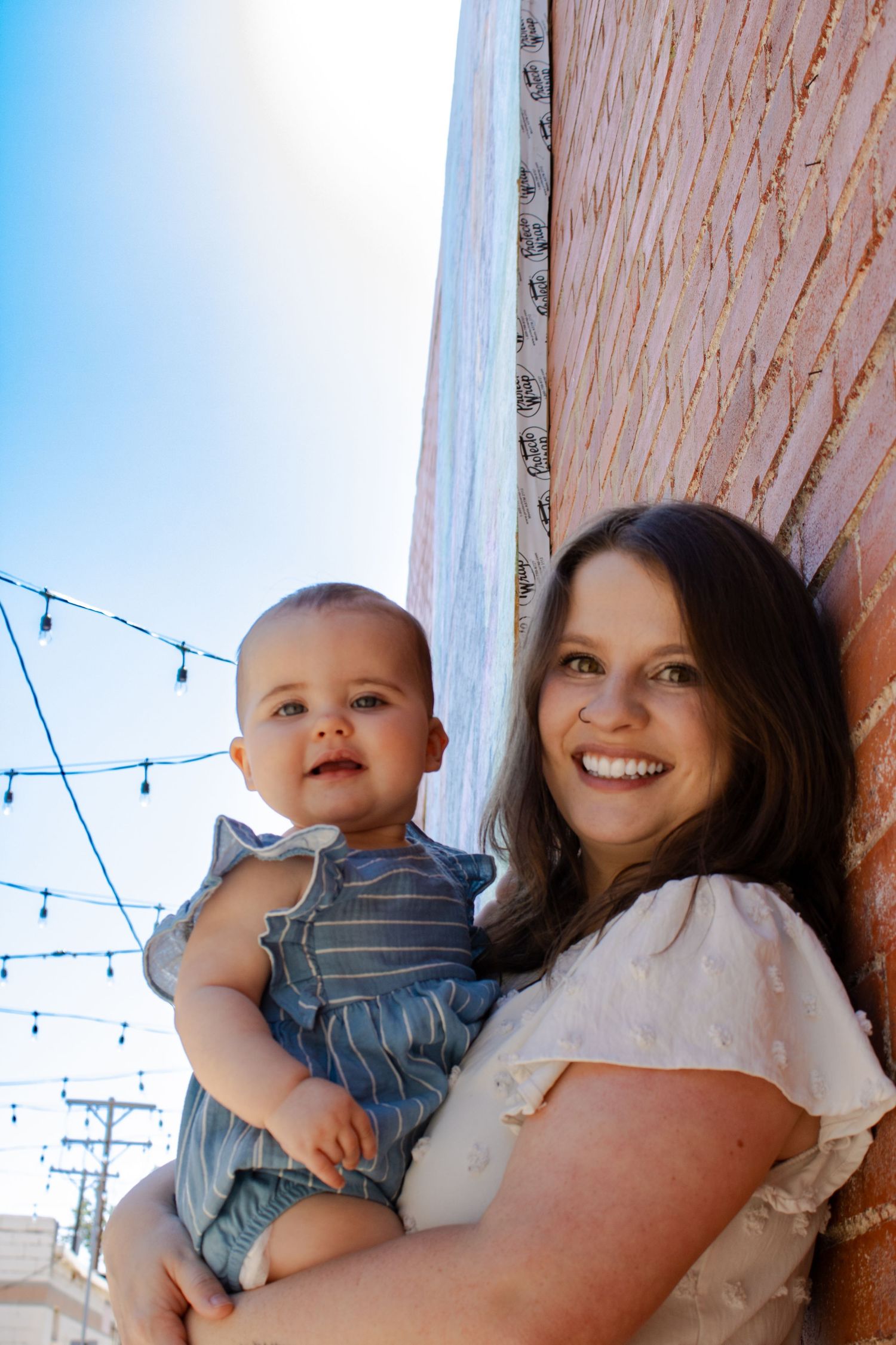 Smiling woman holds a happy baby in a blue dress against a brick wall under blue sky.