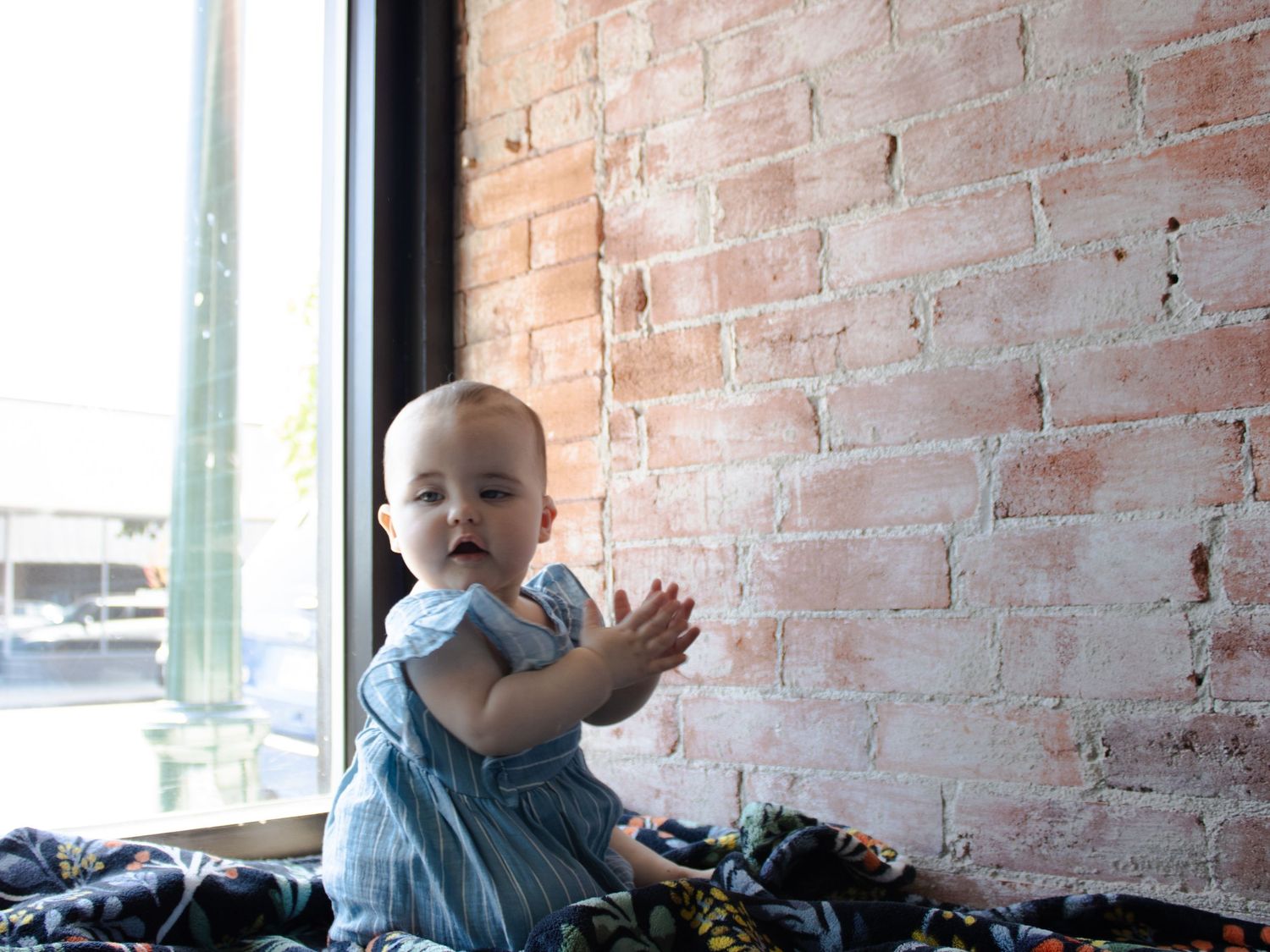 Cute baby in a blue dress clapping hands while sitting near a brick wall by a window.