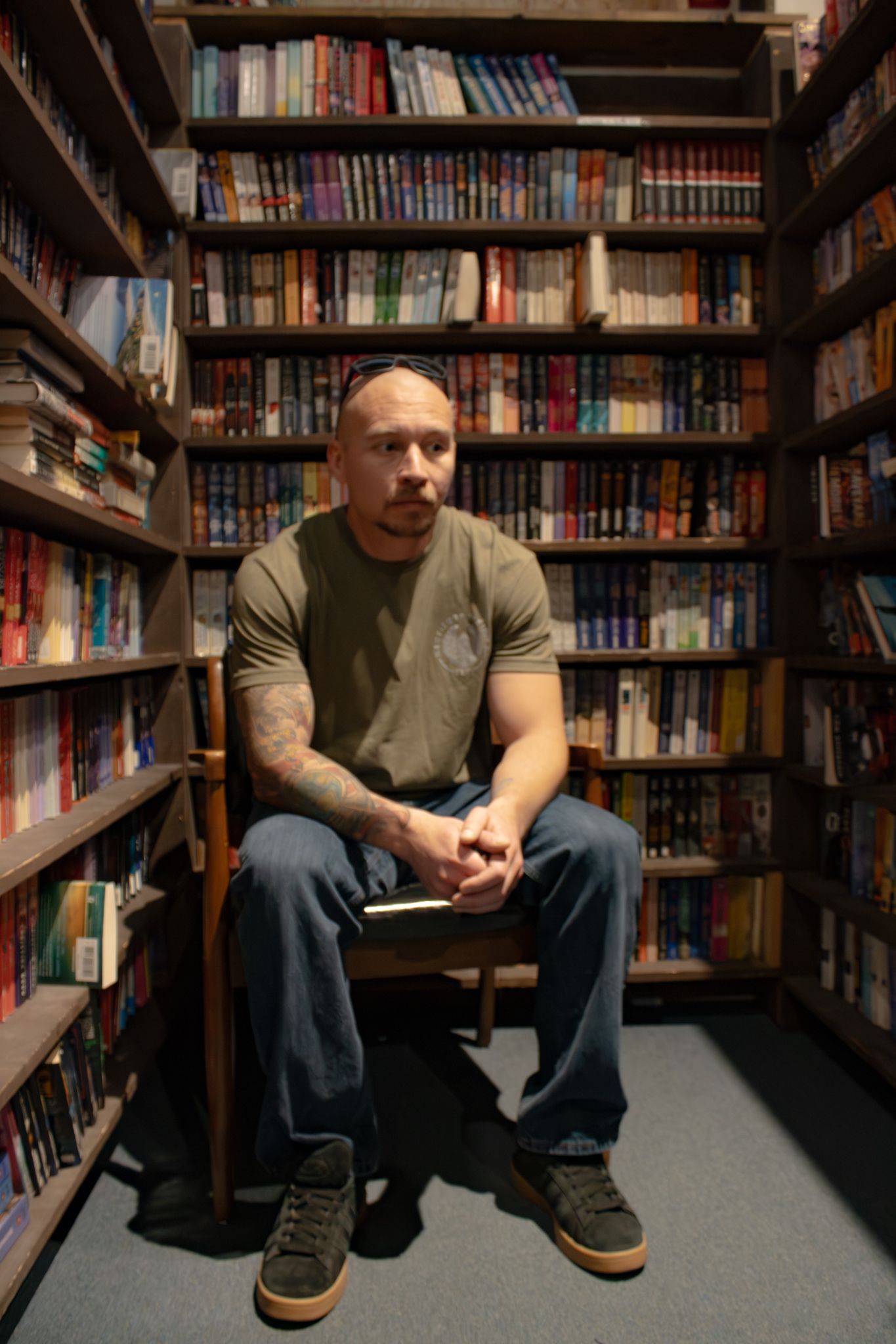 Tattooed man in olive shirt sits on a chair surrounded by tall bookshelves filled with colorful books.