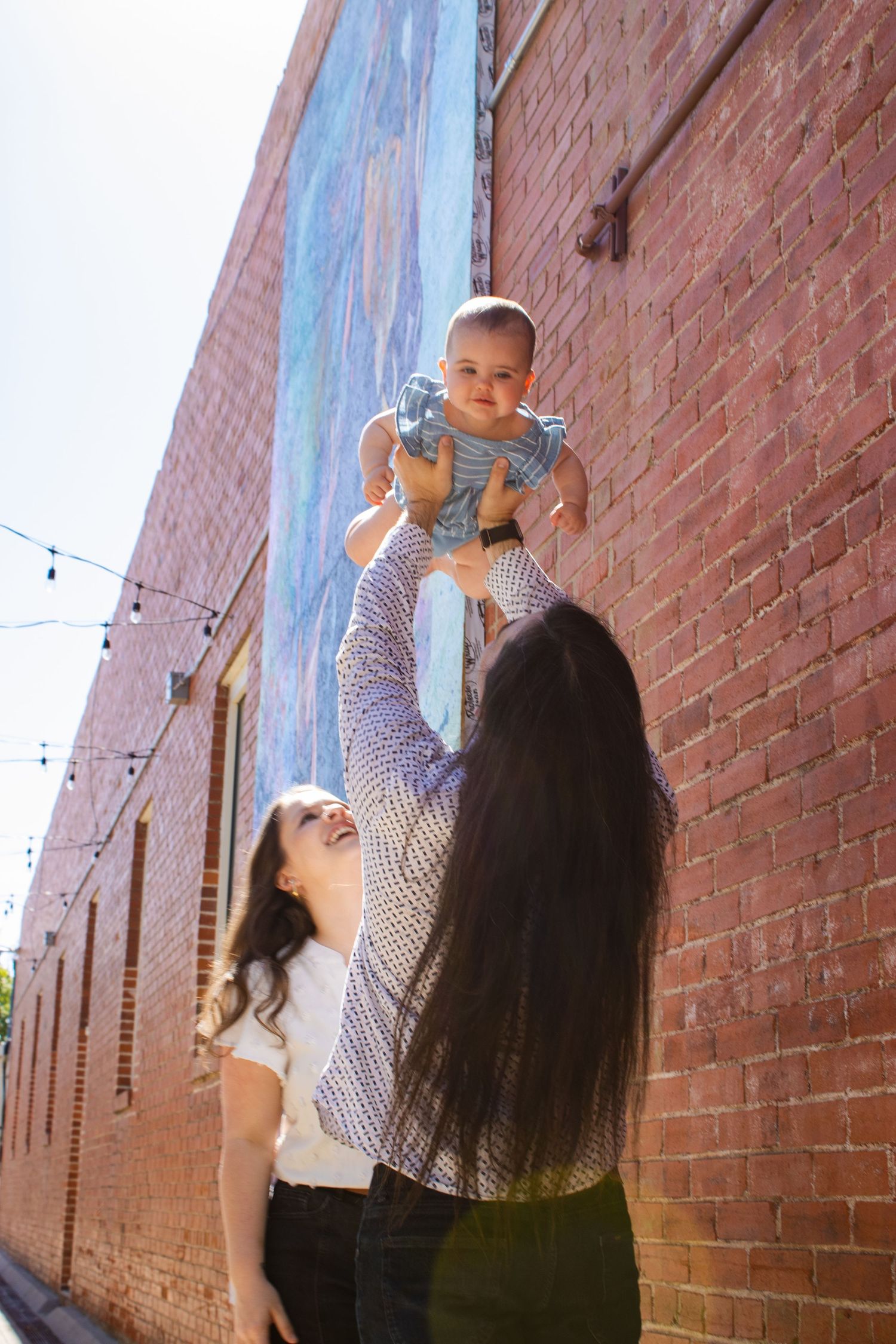 A couple joyfully lift a smiling baby in the air against a brick wall with colorful mural outdoors.