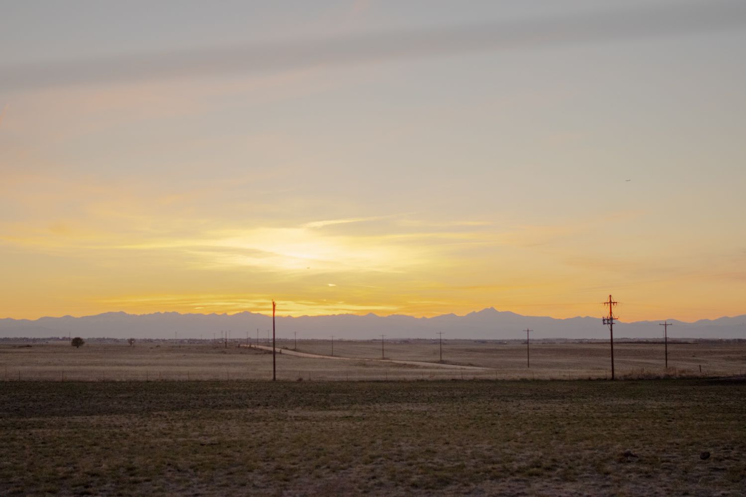 Golden sunset over a flat desert landscape with silhouetted power line poles and distant mountains.