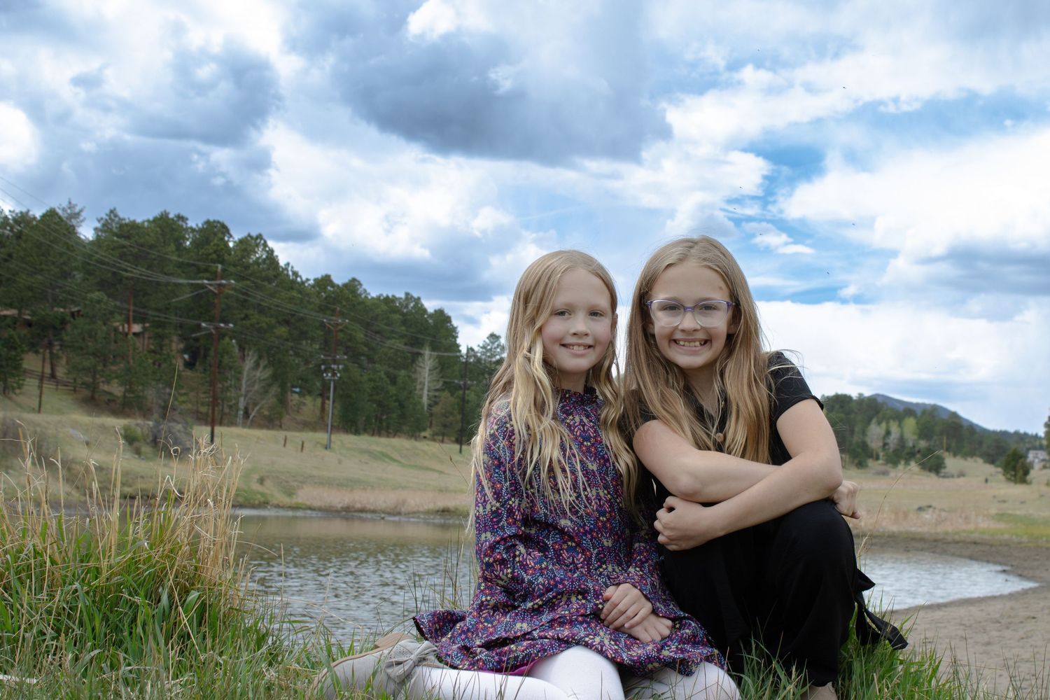 Two young blonde girls smiling and hugging outdoors near a pond with trees and cloudy sky.