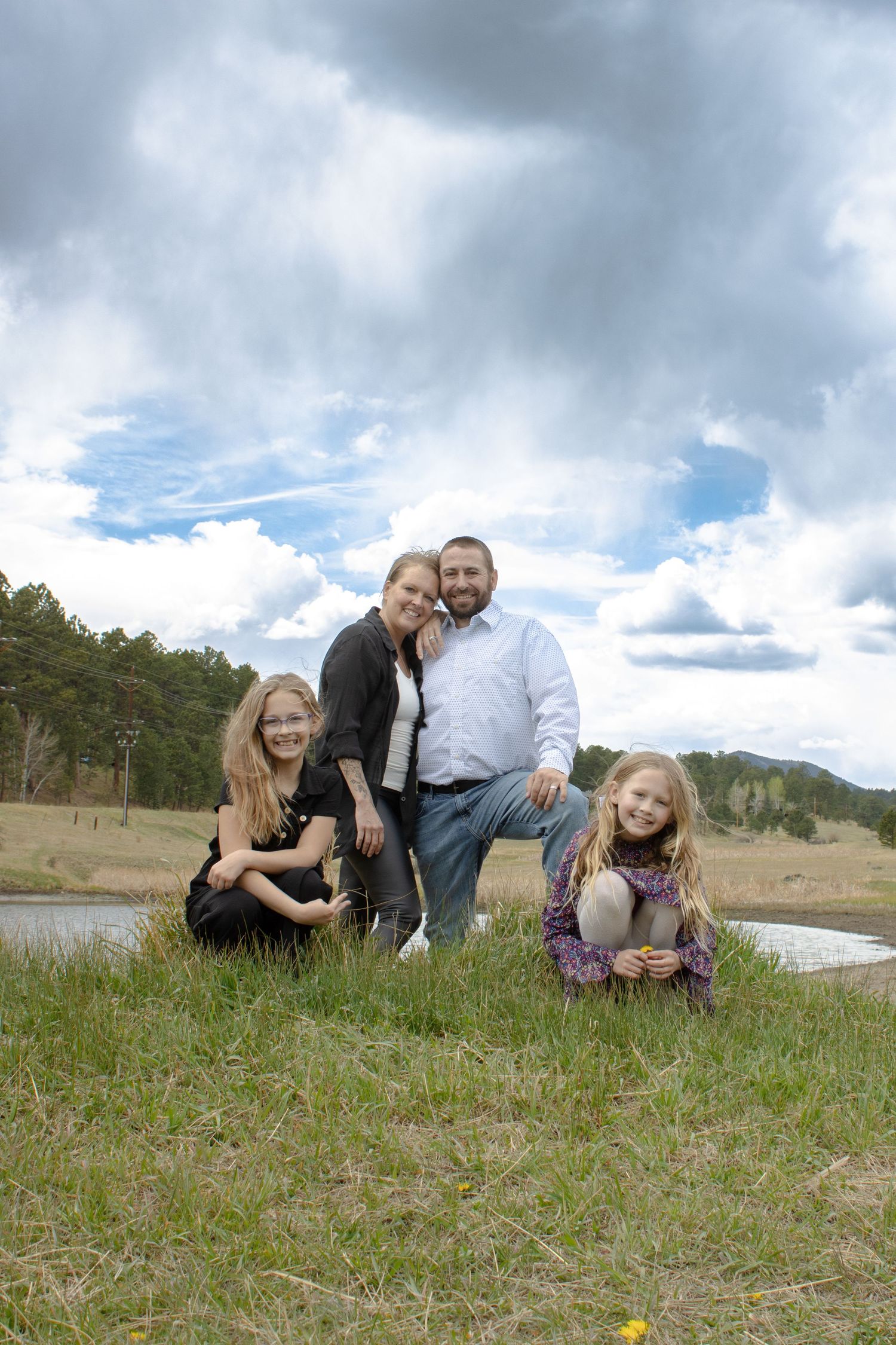Happy family of four poses together in a grassy meadow with a pond and dramatic cloudy sky backdrop.