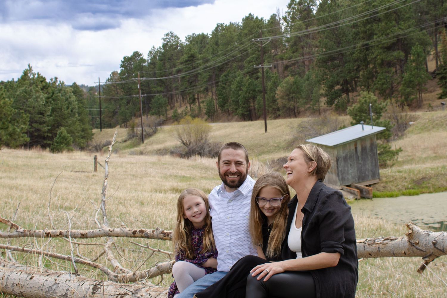 Smiling family of four sits together on a fallen log in a golden mountain meadow under cloudy skies.