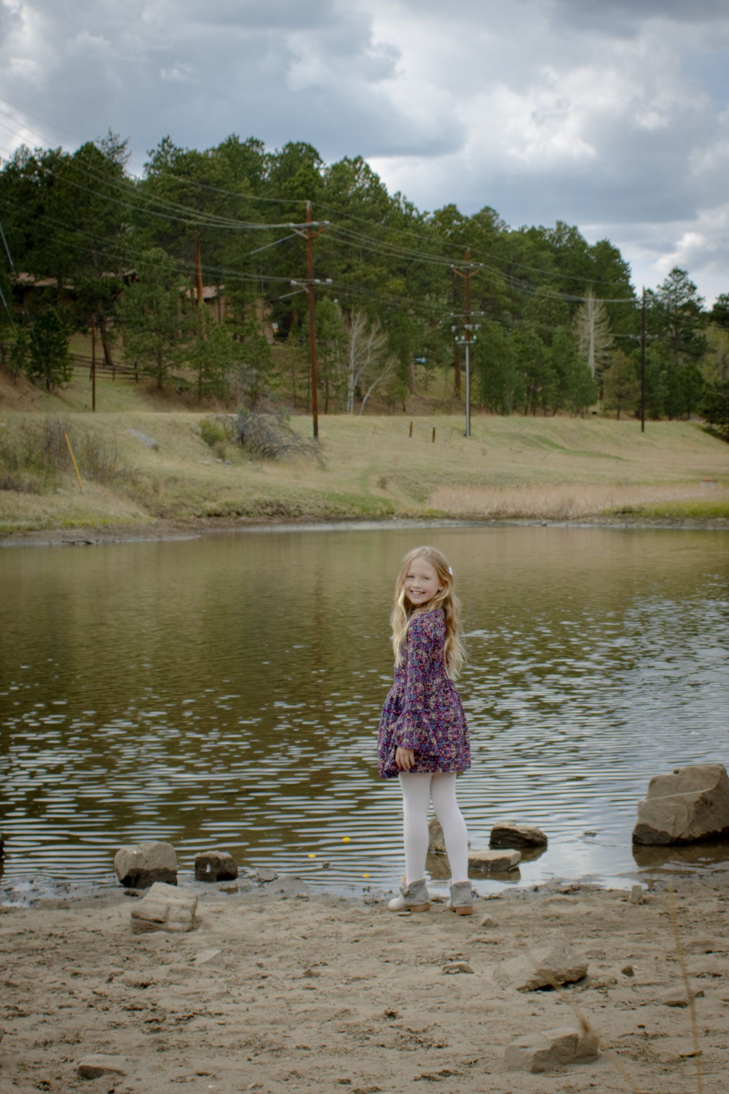Young blonde girl in a purple floral dress standing on rocky shore beside a scenic pond.