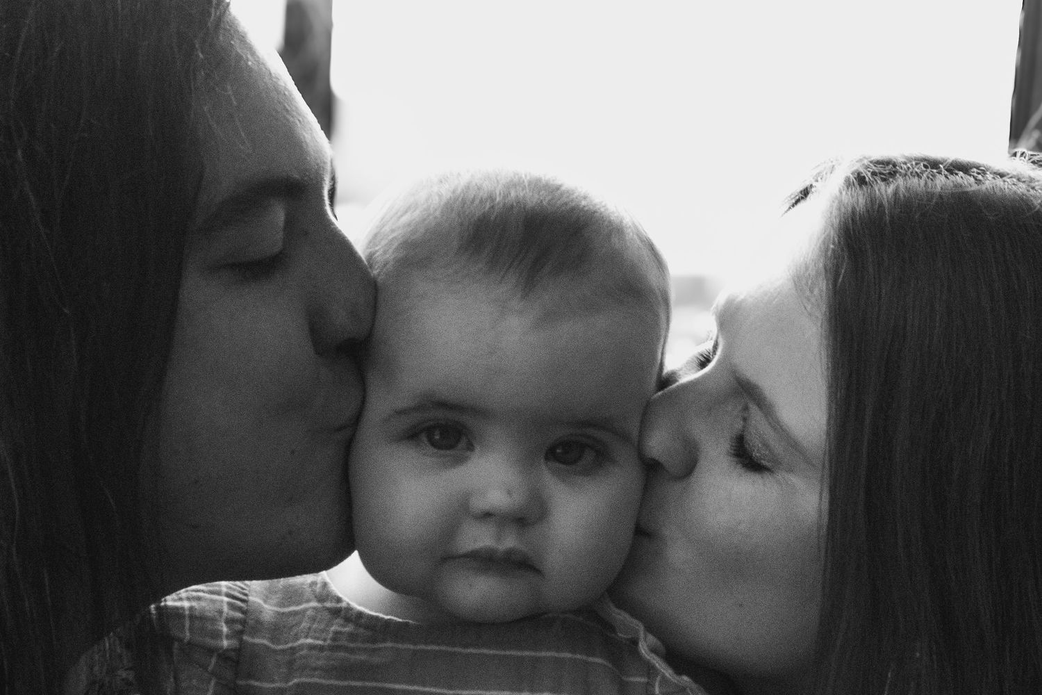 Black and white photo of two adults kissing a baby on the cheeks simultaneously.