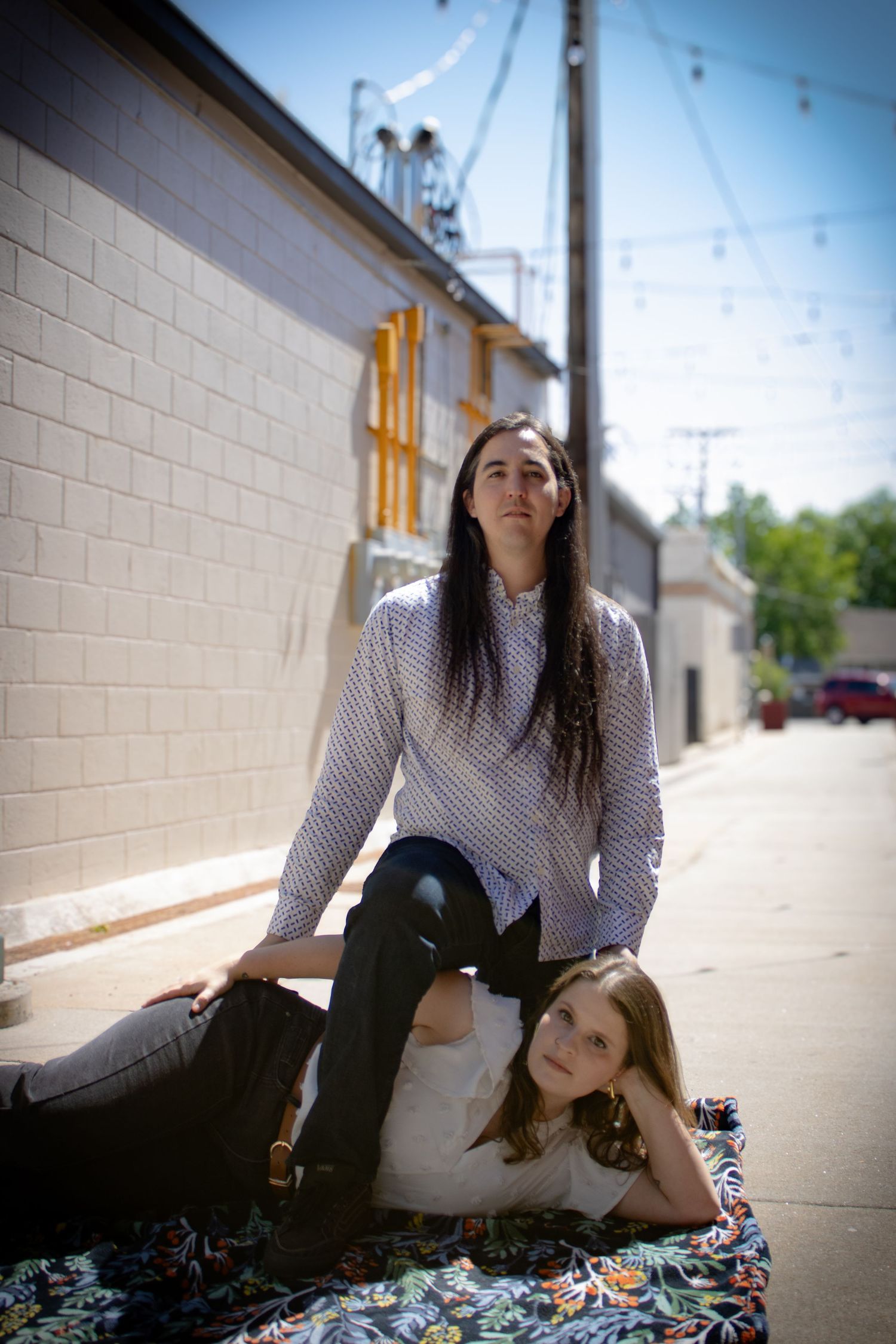 A couple poses in a sunny urban alley, one sitting upright and one reclining on a colorful blanket.