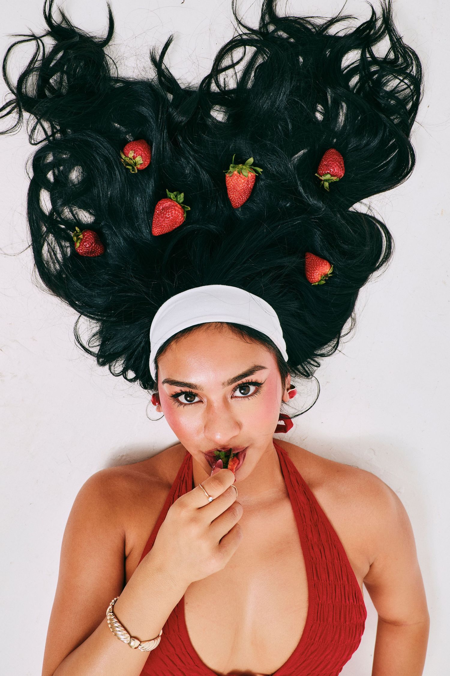 Woman lying down with strawberries in her dark hair eats fruit while wearing a red top.