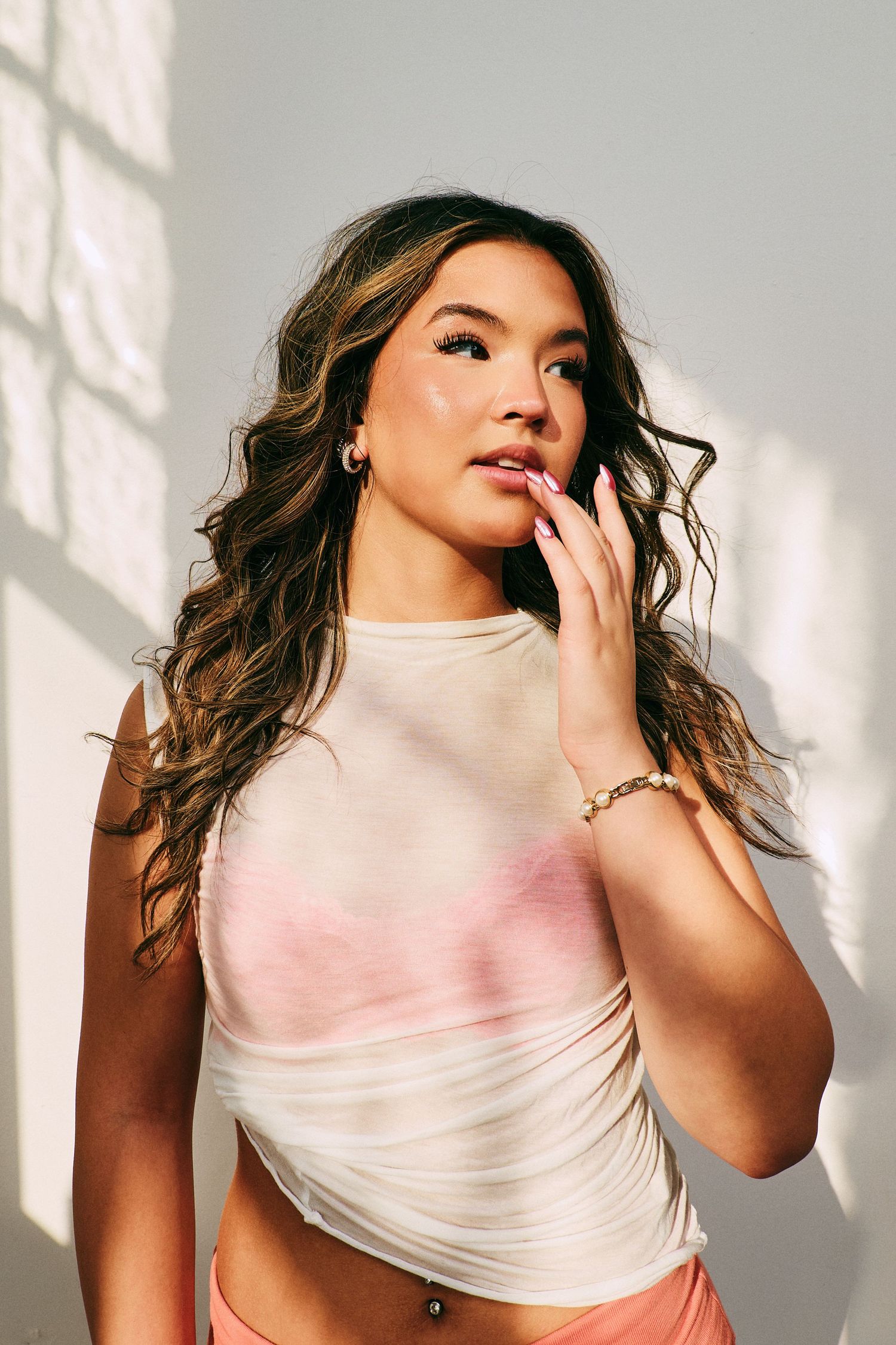 Young woman with wavy hair touches her lips in a sheer white crop top with sunlight shadows.