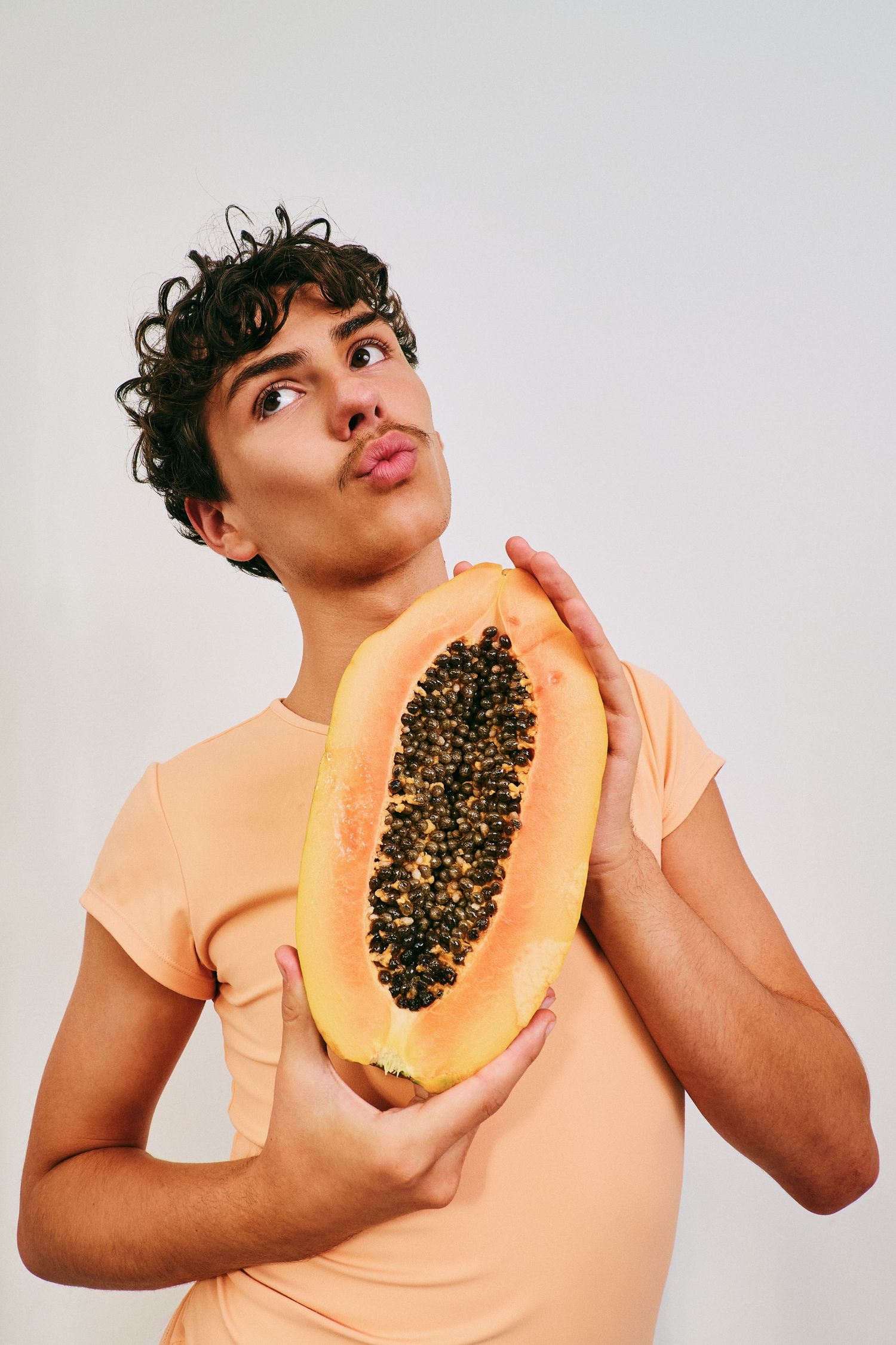 Young man with curly hair holds halved papaya while making a funny face in studio photo.