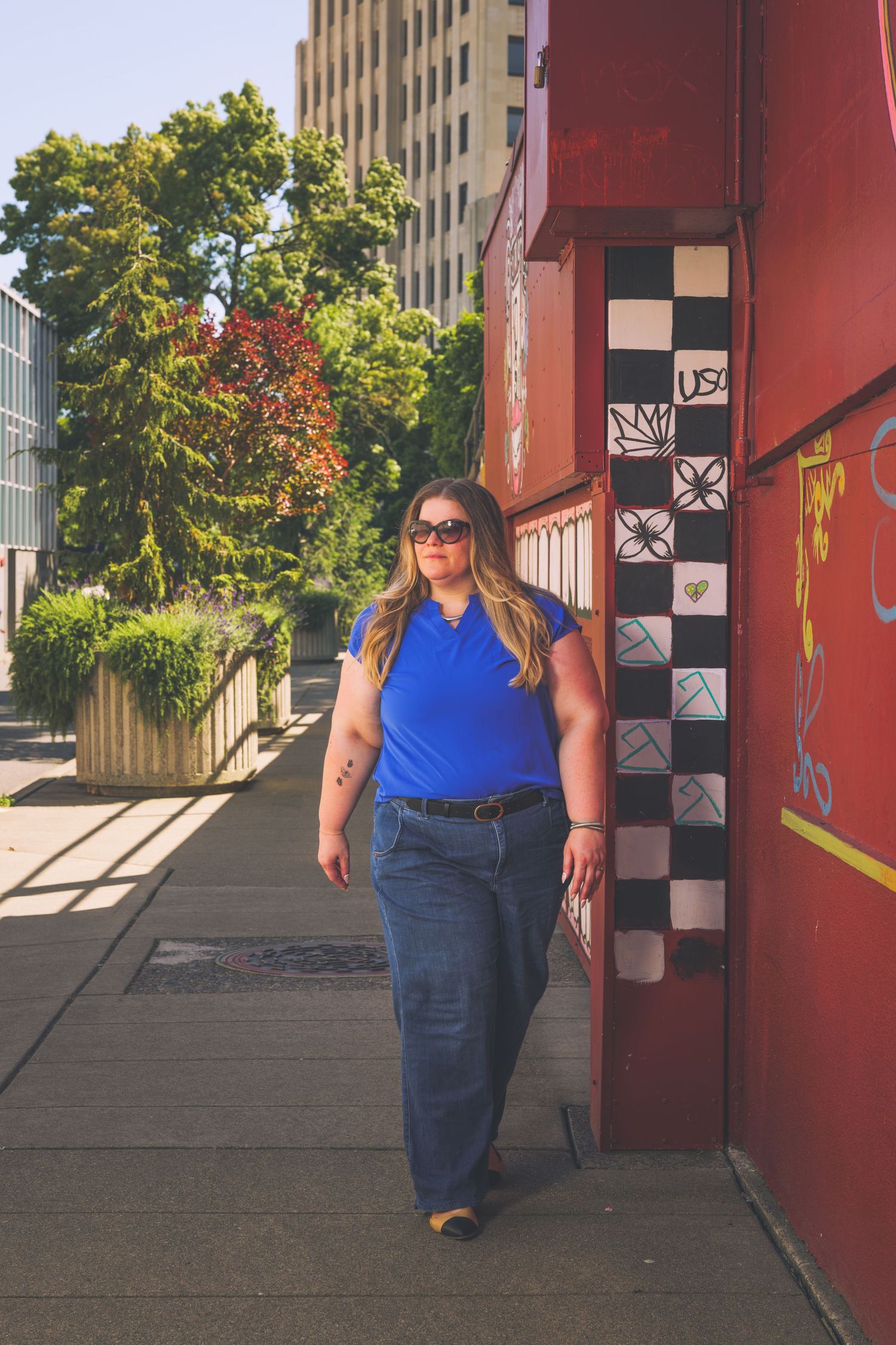 Plus-size woman in blue polo and wide-leg jeans walks confidently on urban sidewalk with murals.