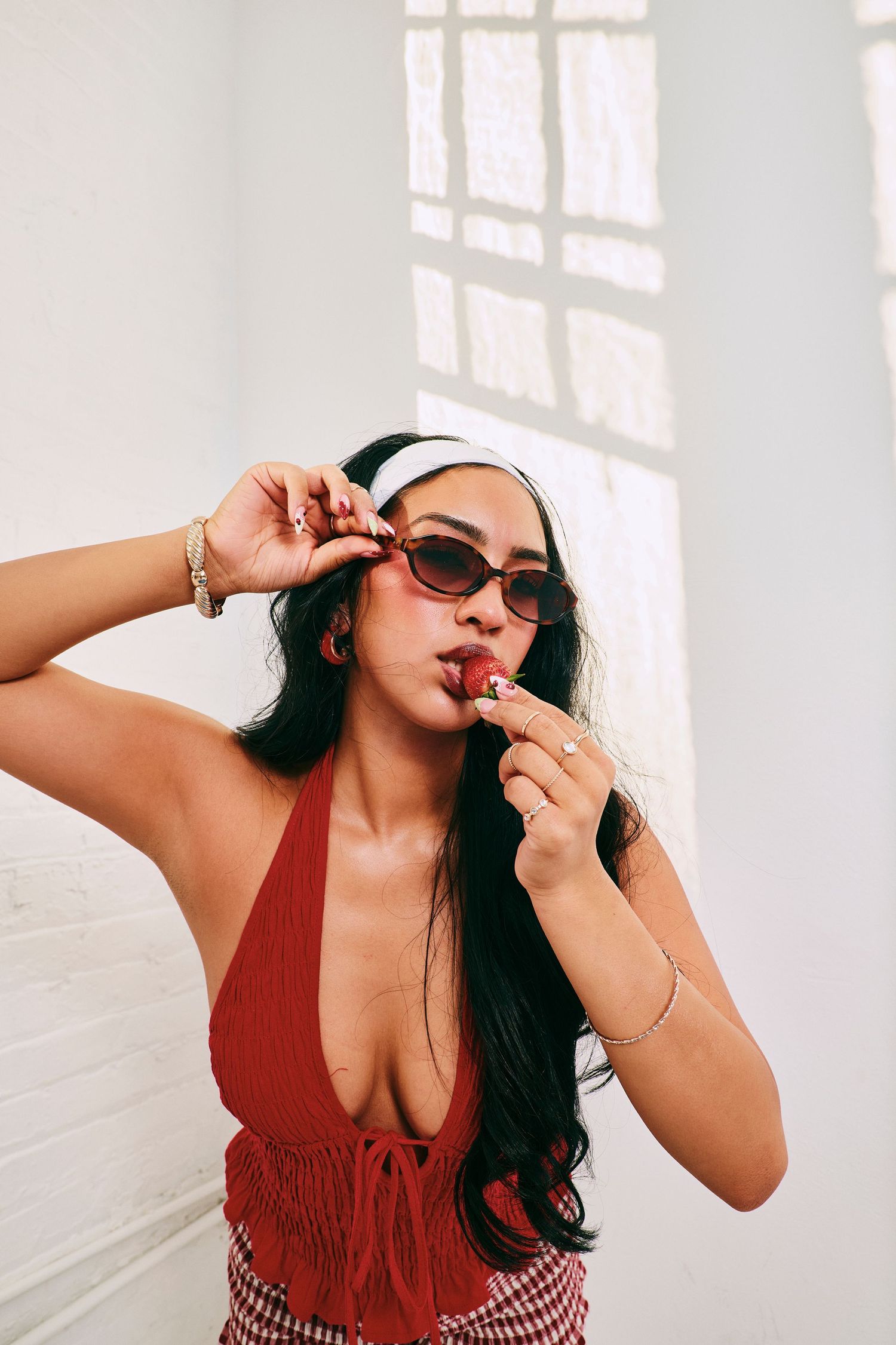 Woman in red halter top and sunglasses eats a strawberry in a bright white room.