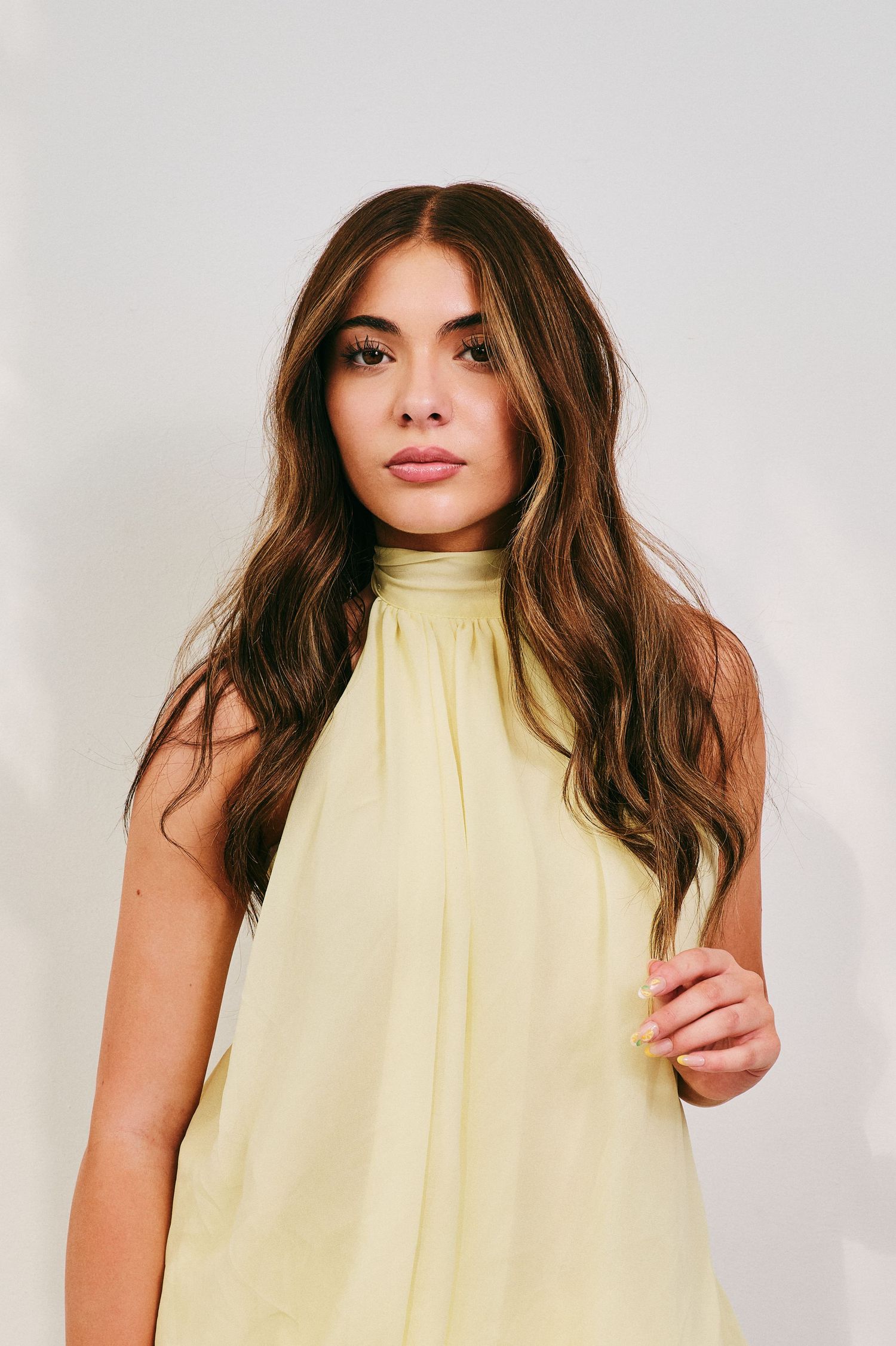 Young woman with long wavy brown hair wears yellow halter top in clean studio portrait.