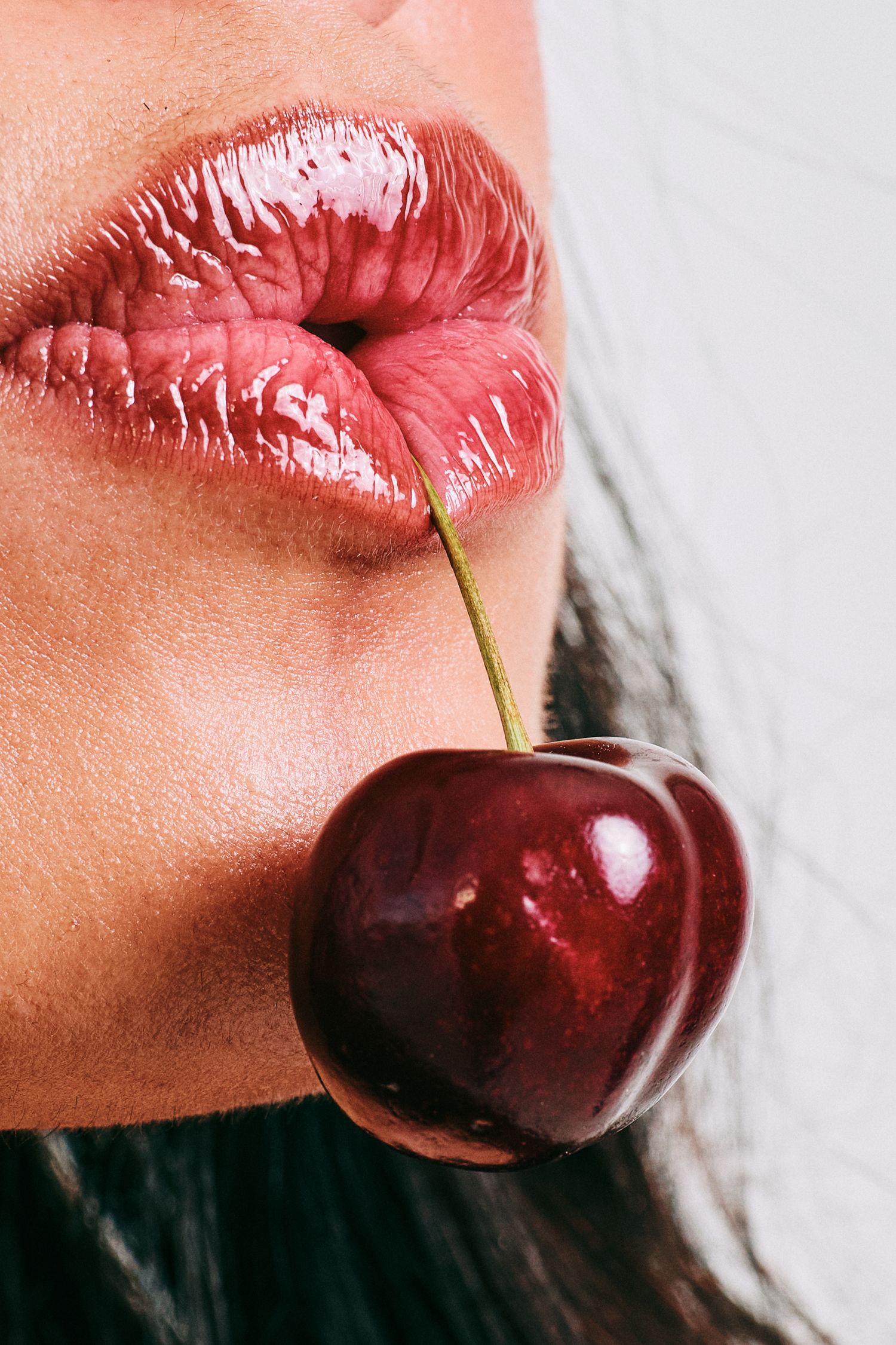 Close-up of glossy pink lips holding a cherry by its stem in a beauty portrait.