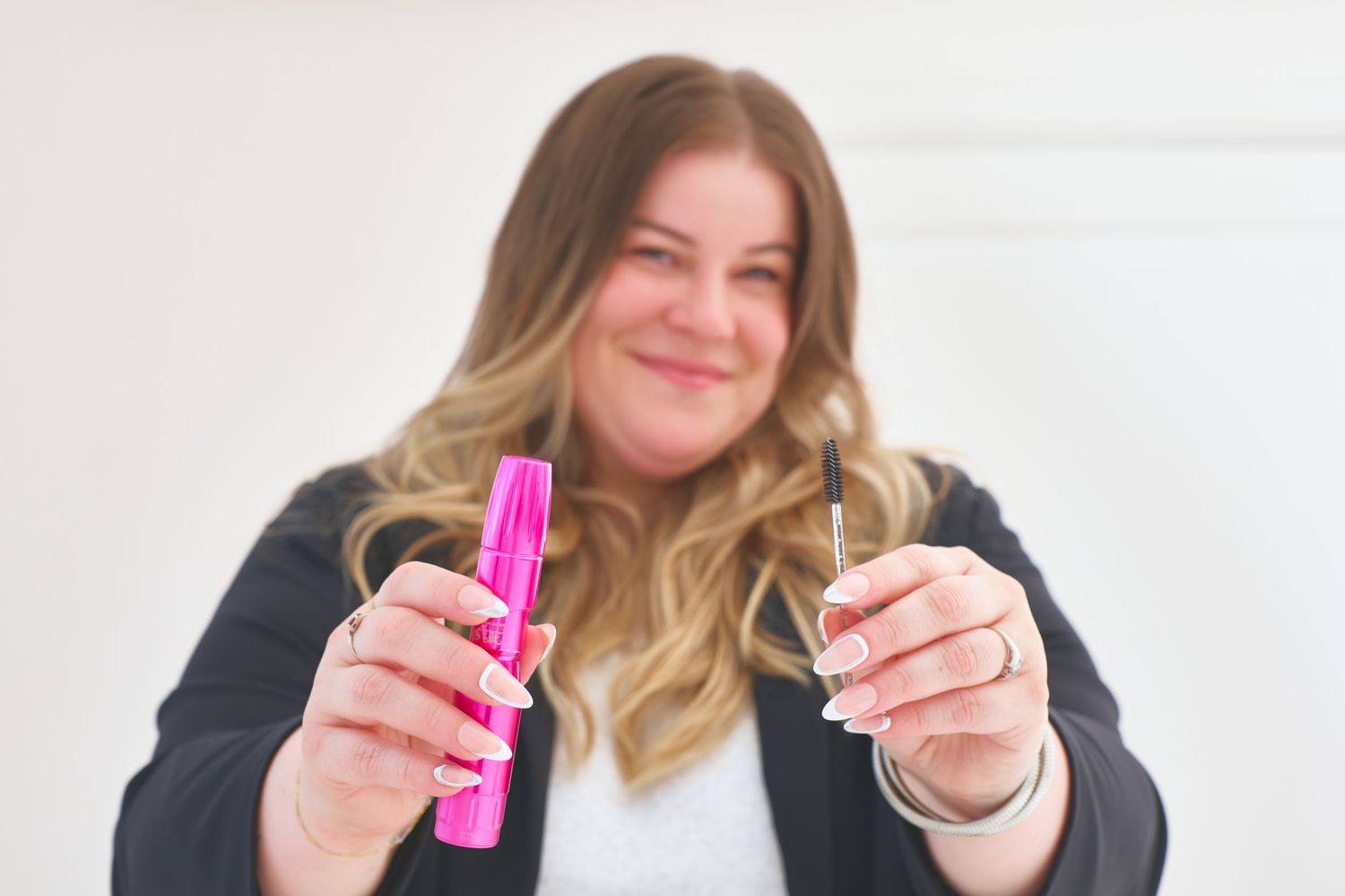 Smiling blonde woman holds pink mascara and eyeliner toward camera in bright studio setting.