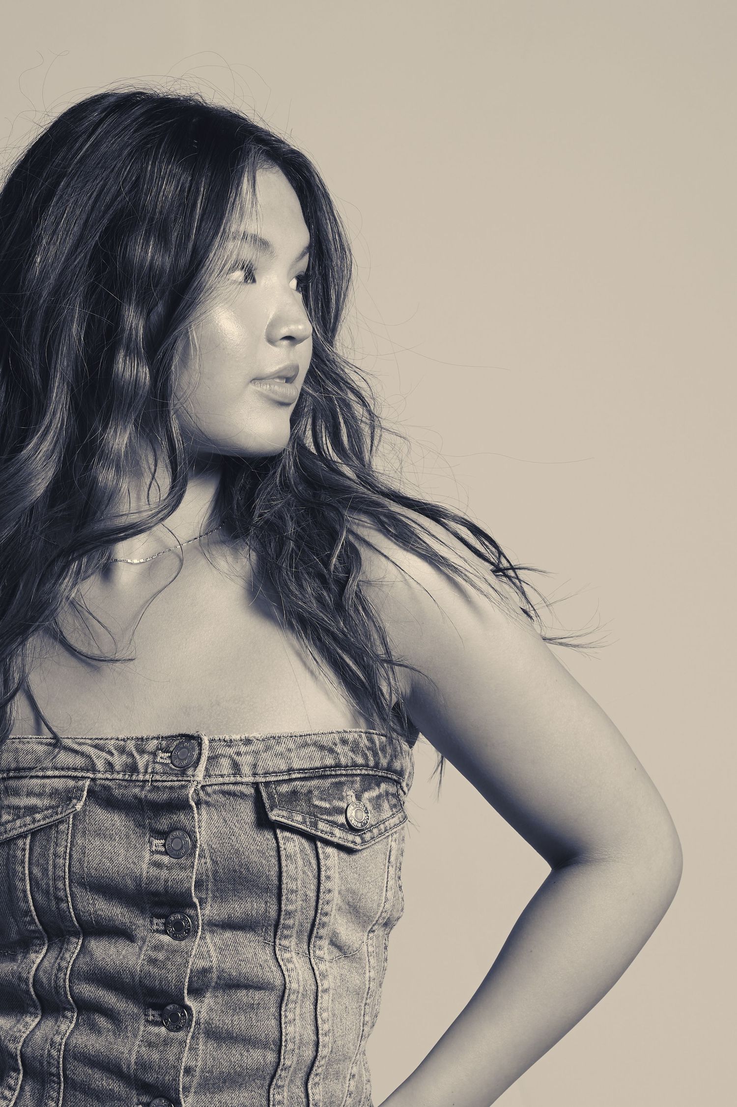 Young Asian woman with long wavy hair wearing a denim jumpsuit poses in a black and white studio portrait.