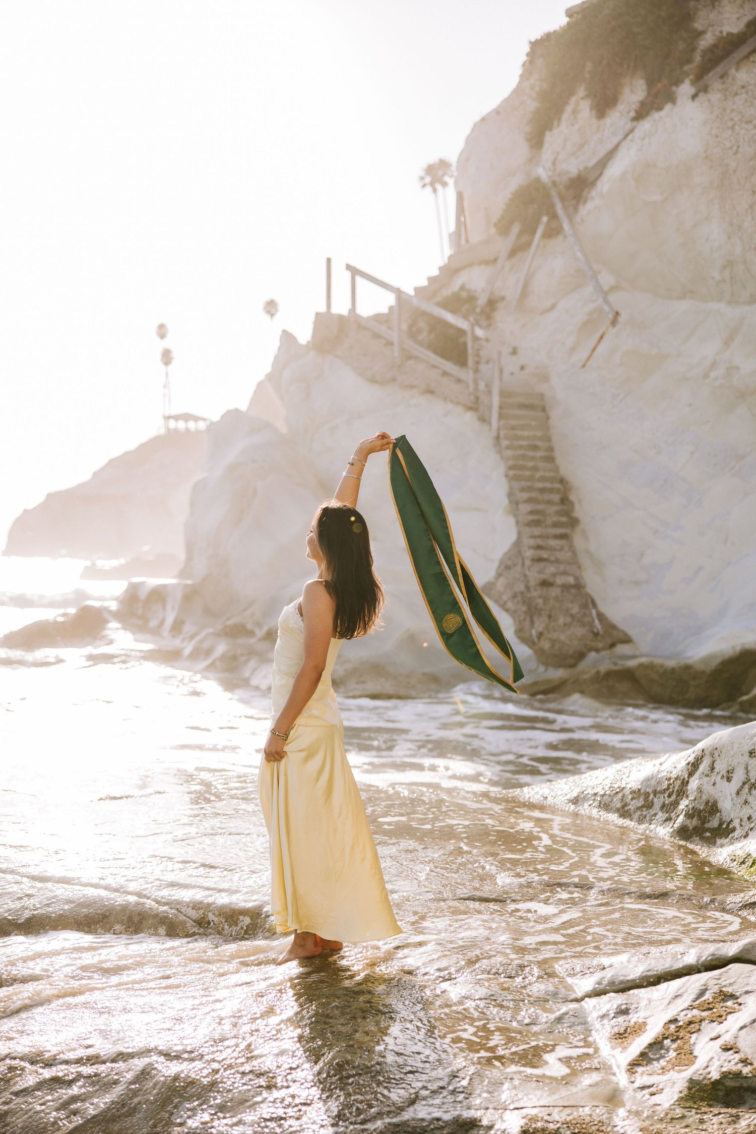 Woman in yellow maxi dress raising green graduation stole at rocky beach during sunset.