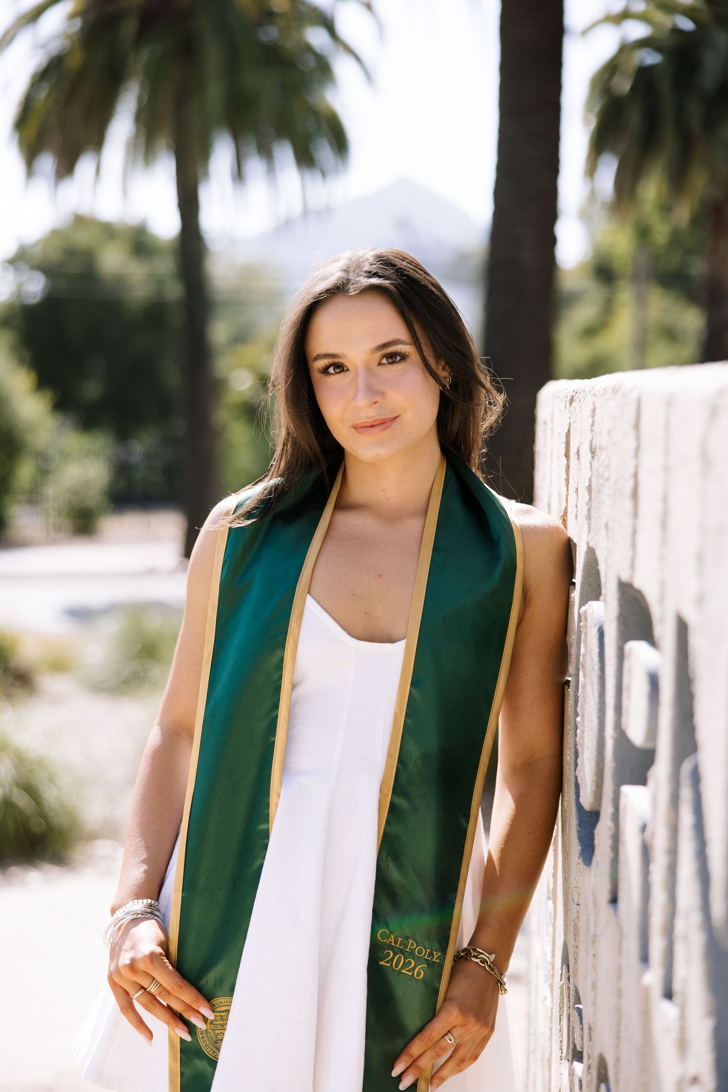 Brunette woman in white dress with Cal Poly 2026 green graduation stole leaning on wall outdoors.