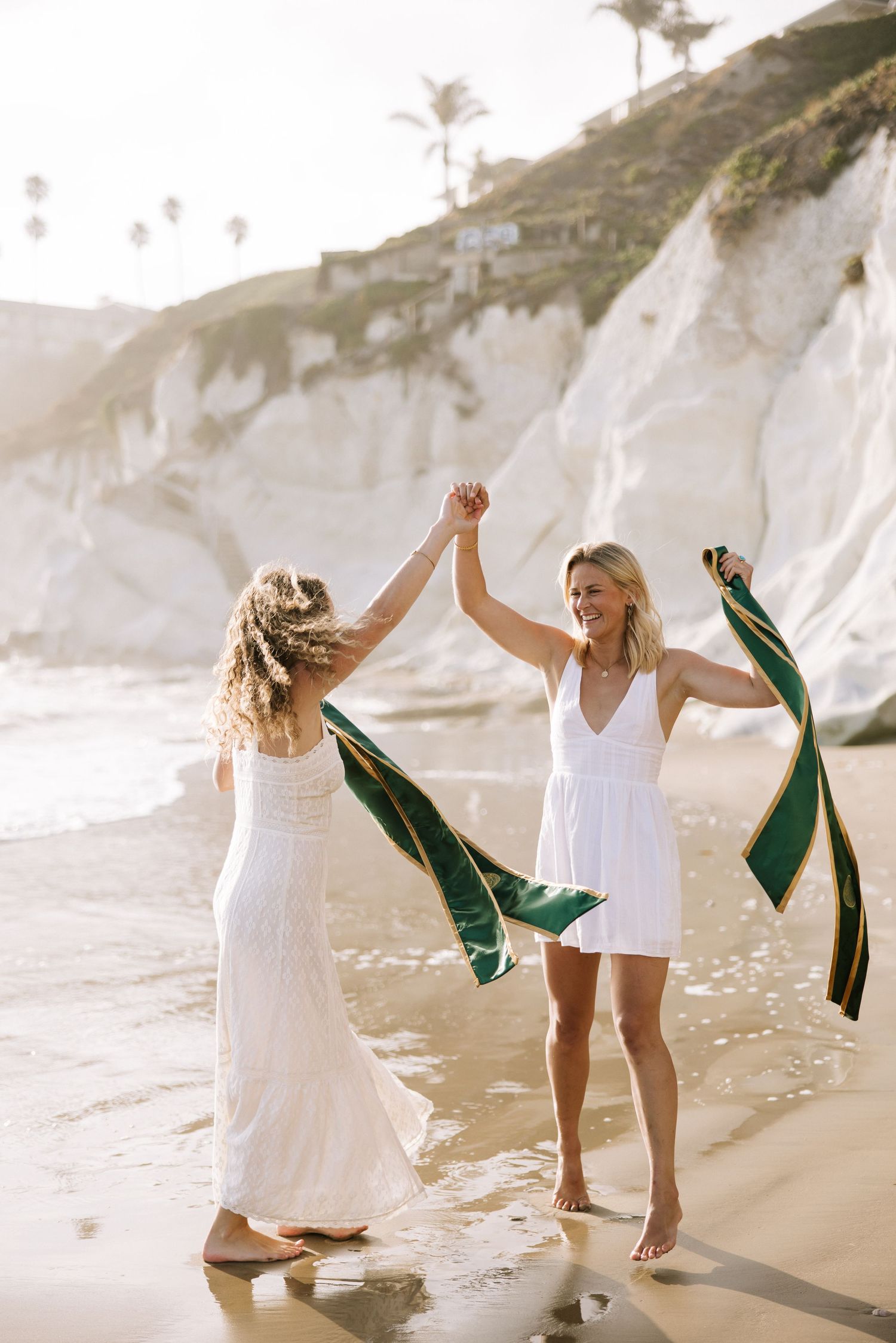 Two women in white dresses dance on a sunny beach holding a green graduation sash.