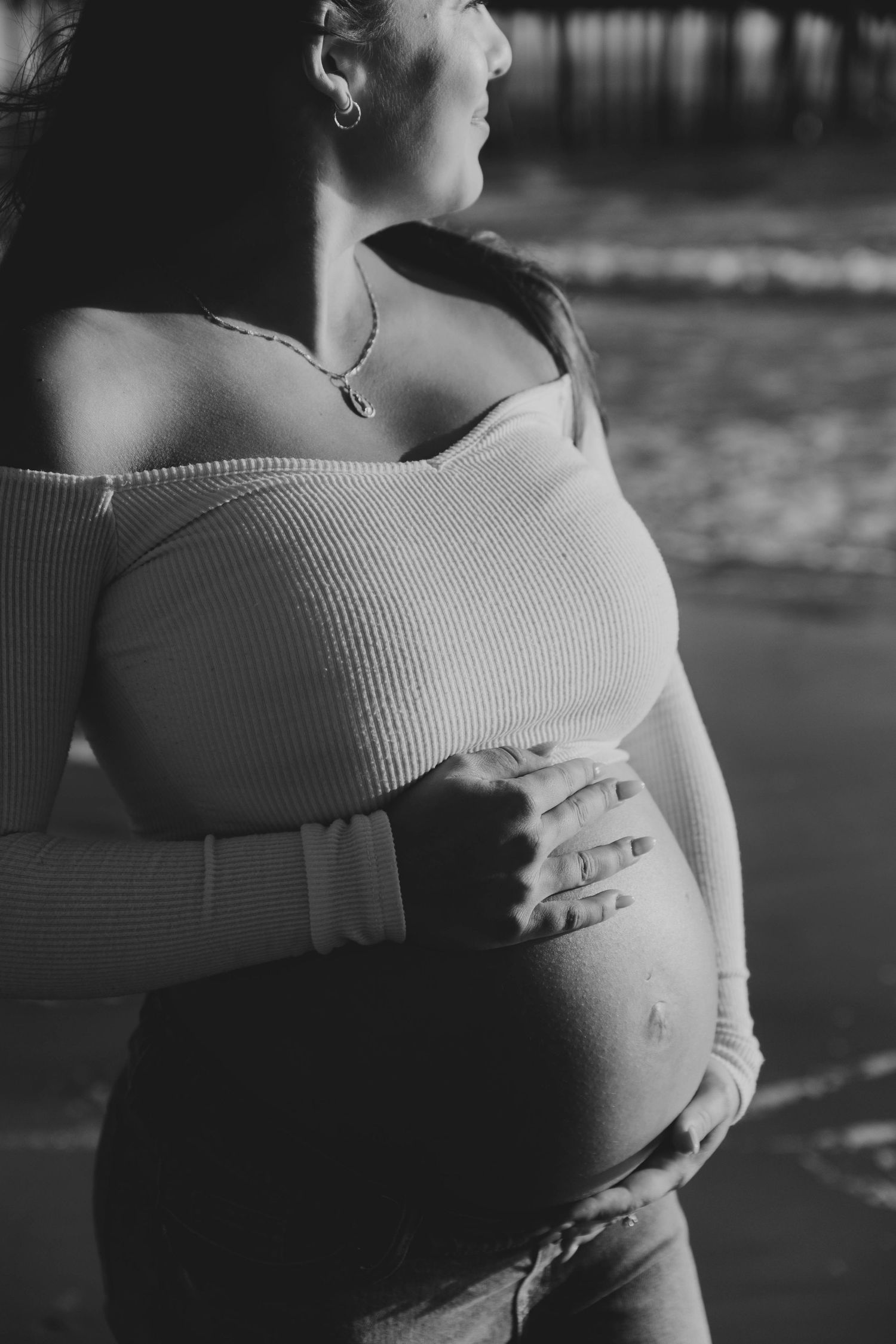 Black and white artistic maternity photo of a pregnant woman in an off-shoulder top holding her belly near a pier.