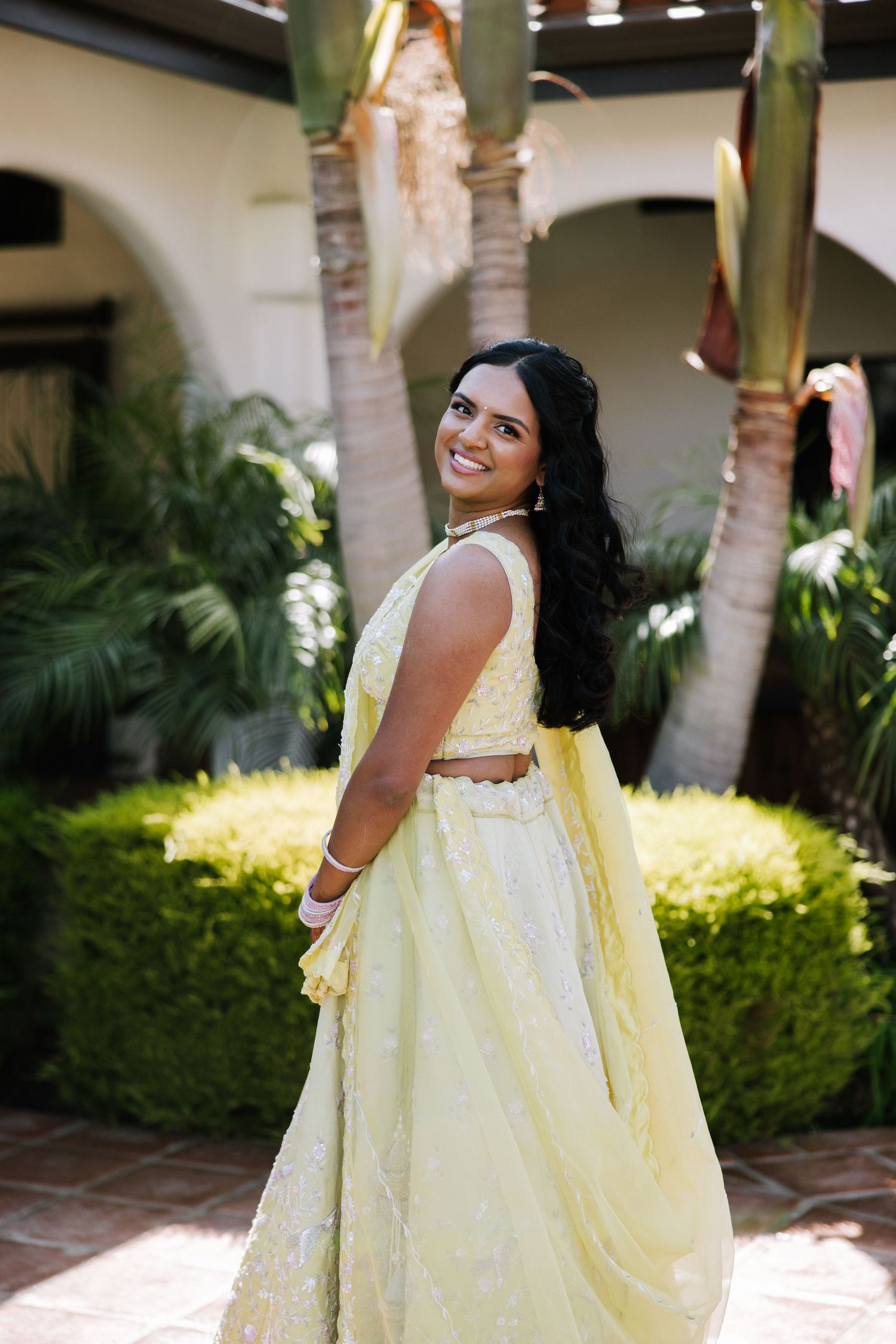 Indian bride in yellow embroidered lehenga smiles over shoulder in tropical garden setting.