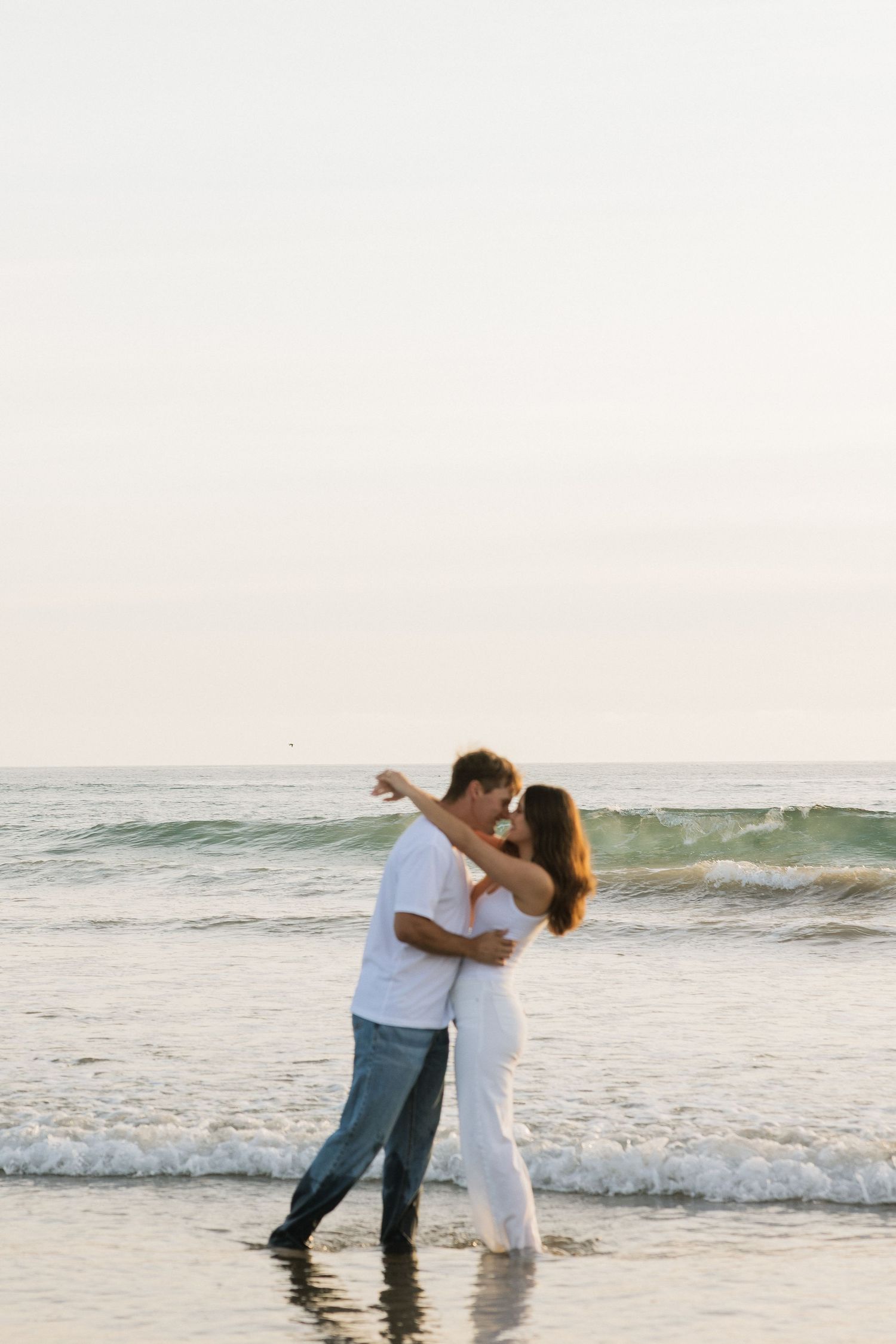 Couple embracing and almost kissing while standing in ocean waves at sunset beach.