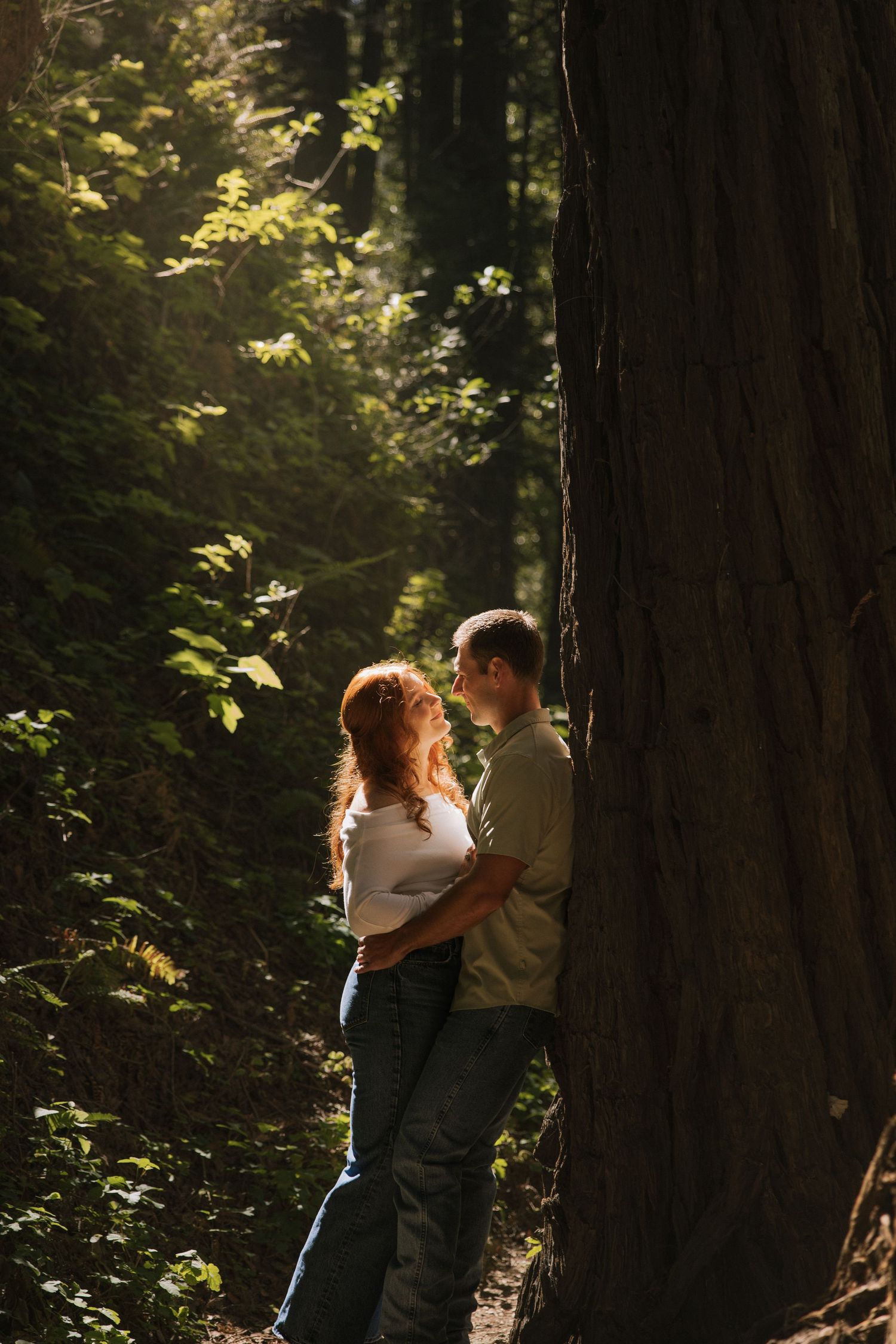 Couple embraces romantically beside a large redwood tree in a sunlit forest.