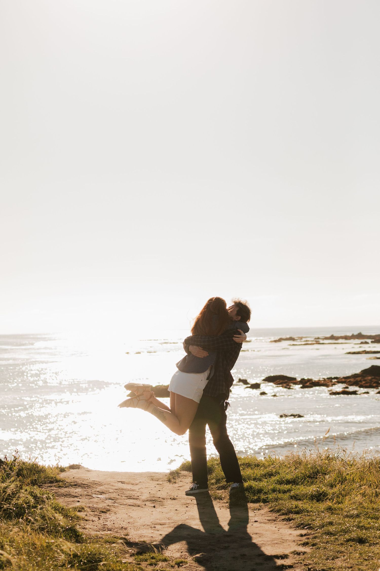 Man lifting woman in joyful embrace on coastal path with sparkling ocean backdrop at sunset.