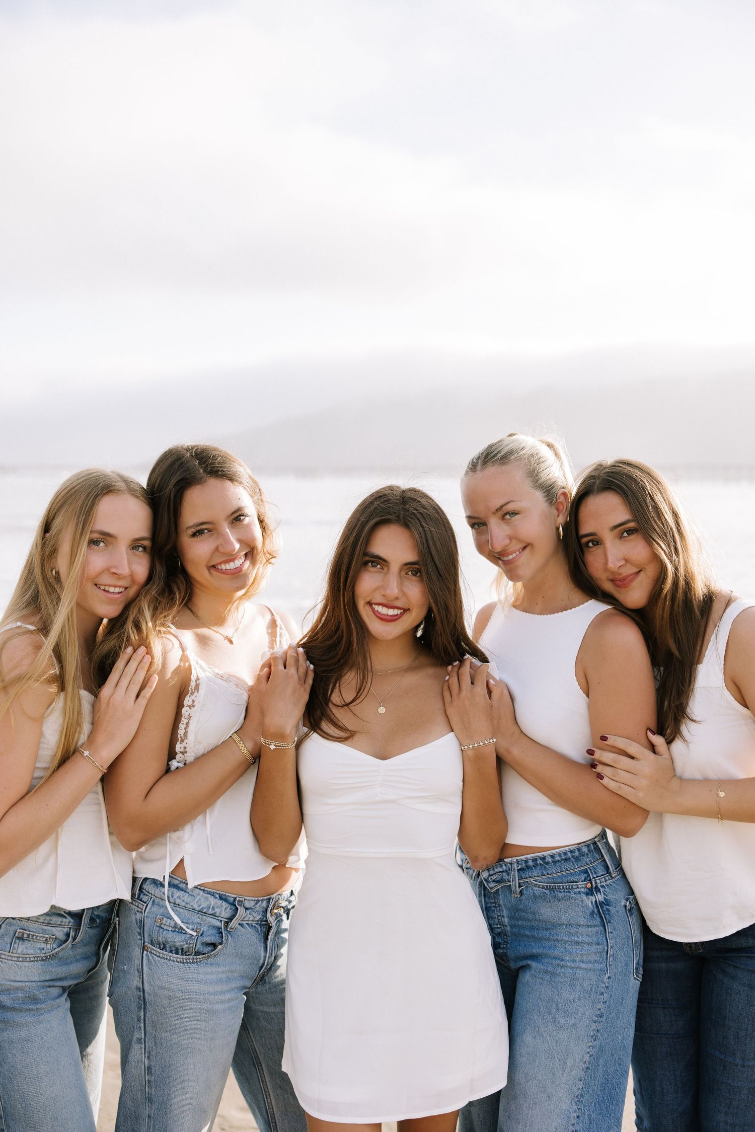 Five young women in white tops and jeans pose together near ocean with overcast sky backdrop.