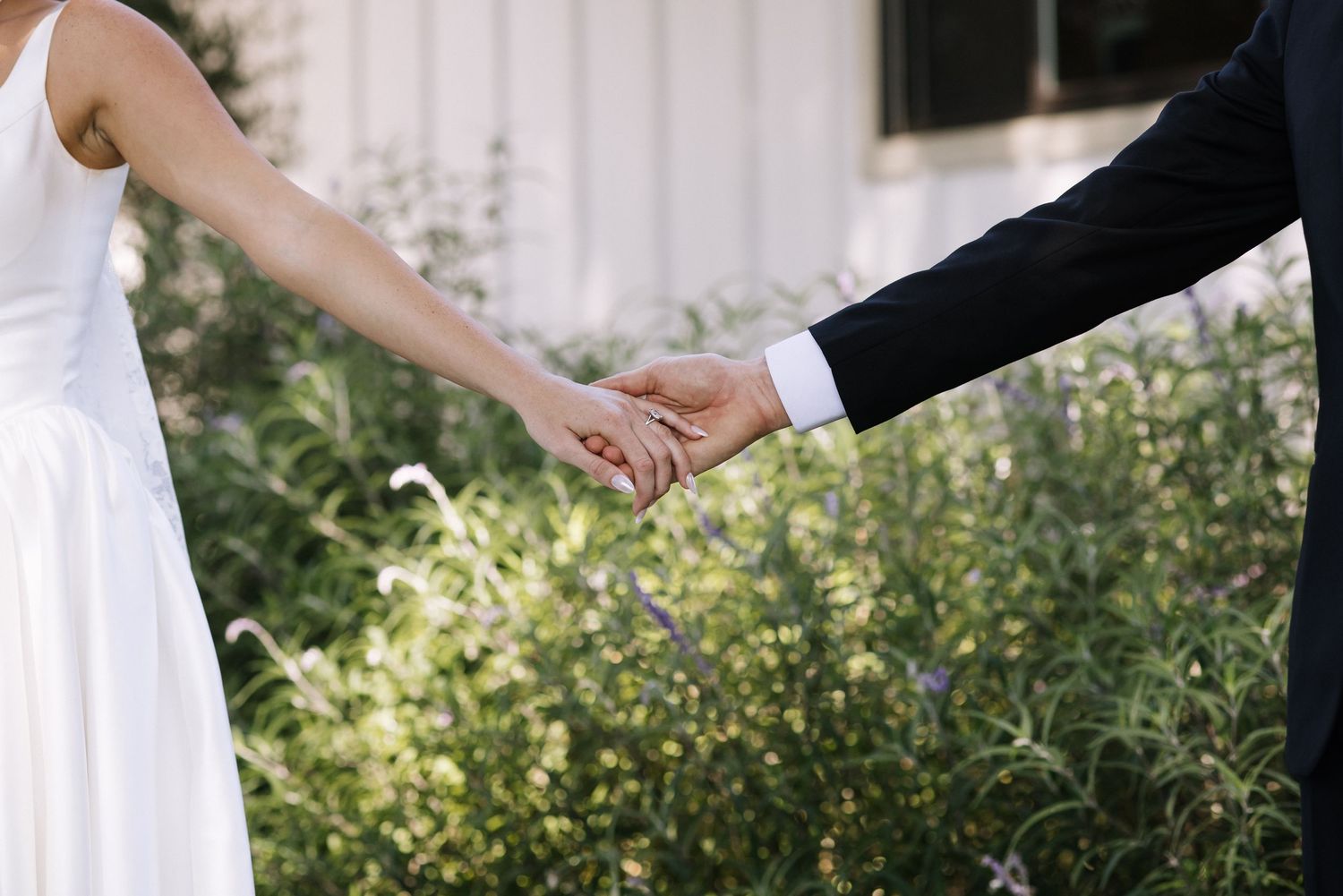 A bride and groom hold hands outdoors near lush greenery, wearing a white wedding dress and black suit.