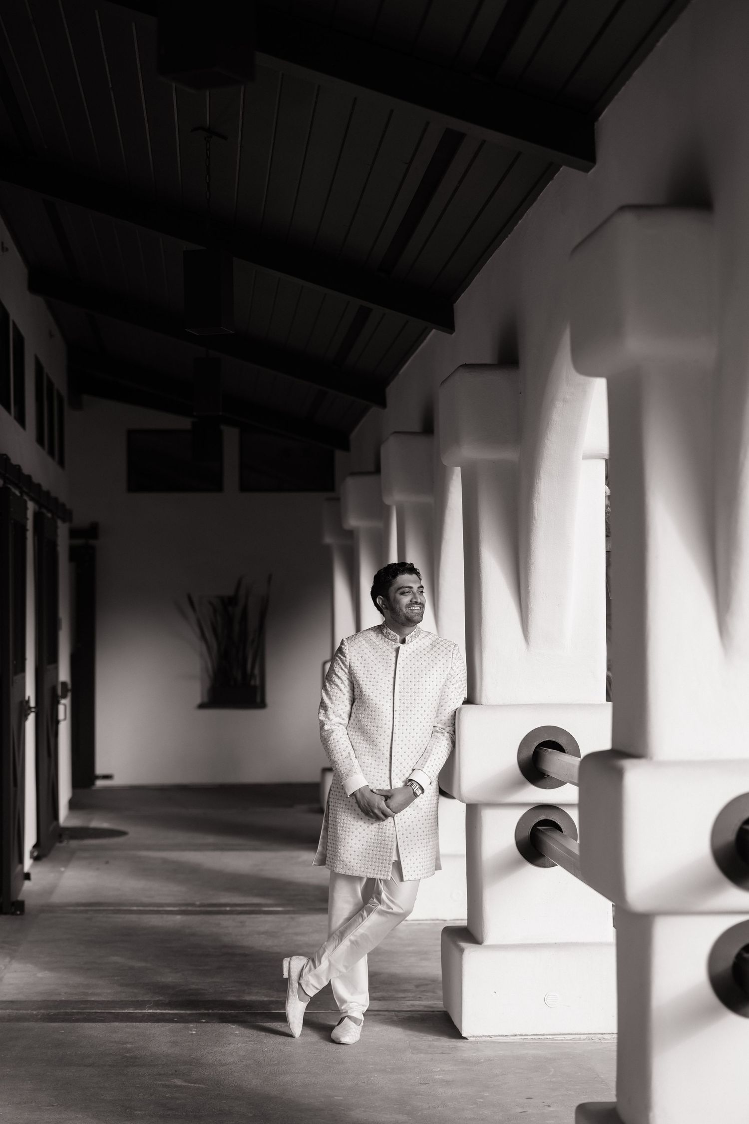Groom in white sherwani posing elegantly in a black and white architectural corridor.