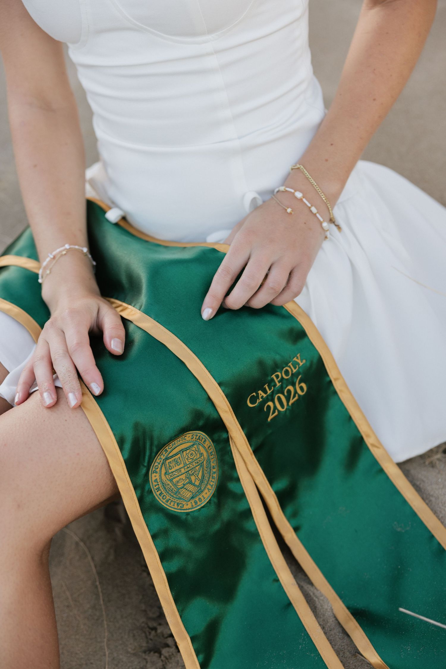 Close-up of Cal Poly 2026 green graduation sash on a woman in white dress with gold bracelet.