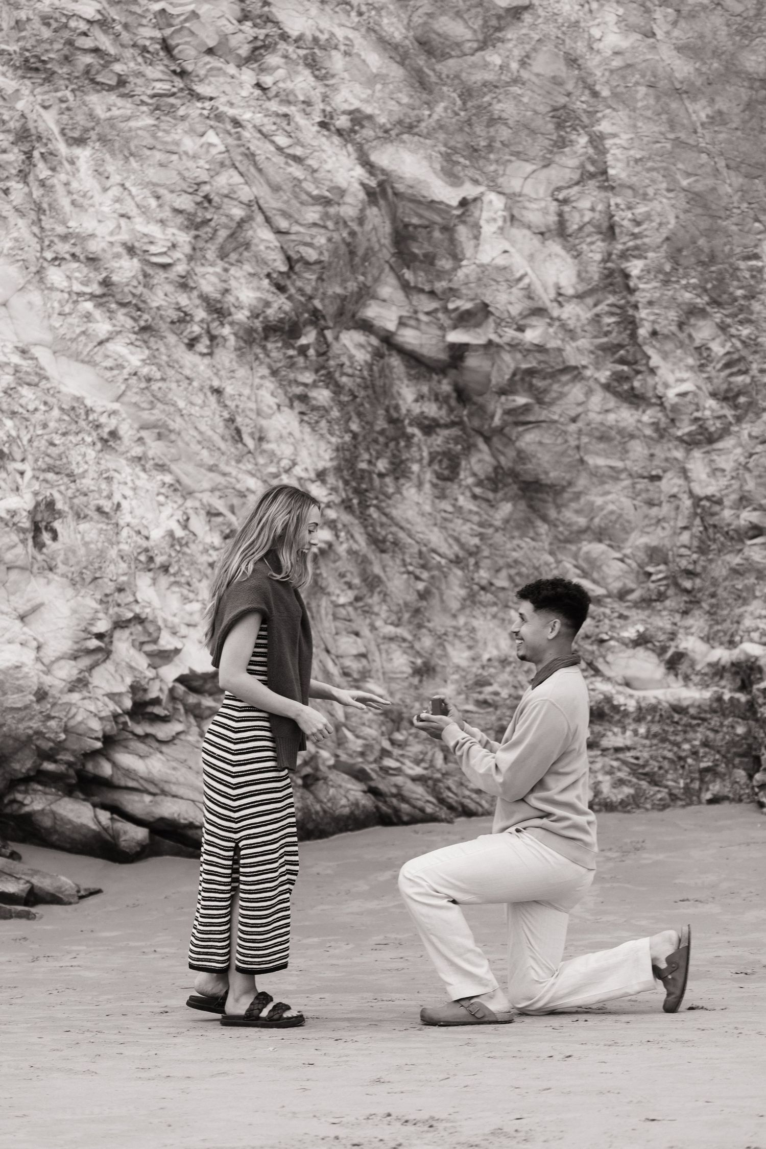Black and white photo of a man on one knee proposing to a woman on a sandy beach by rock cliffs.