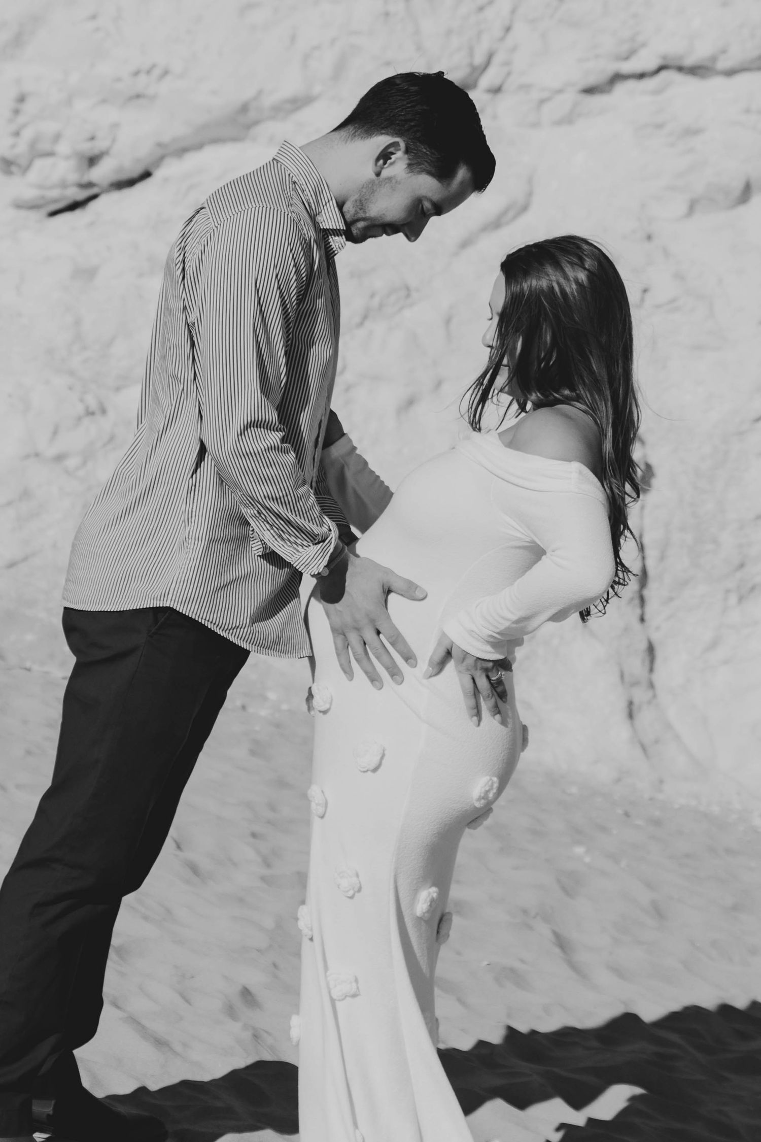 Black and white photo of couple on sandy beach, man tenderly touching pregnant womans baby bump.