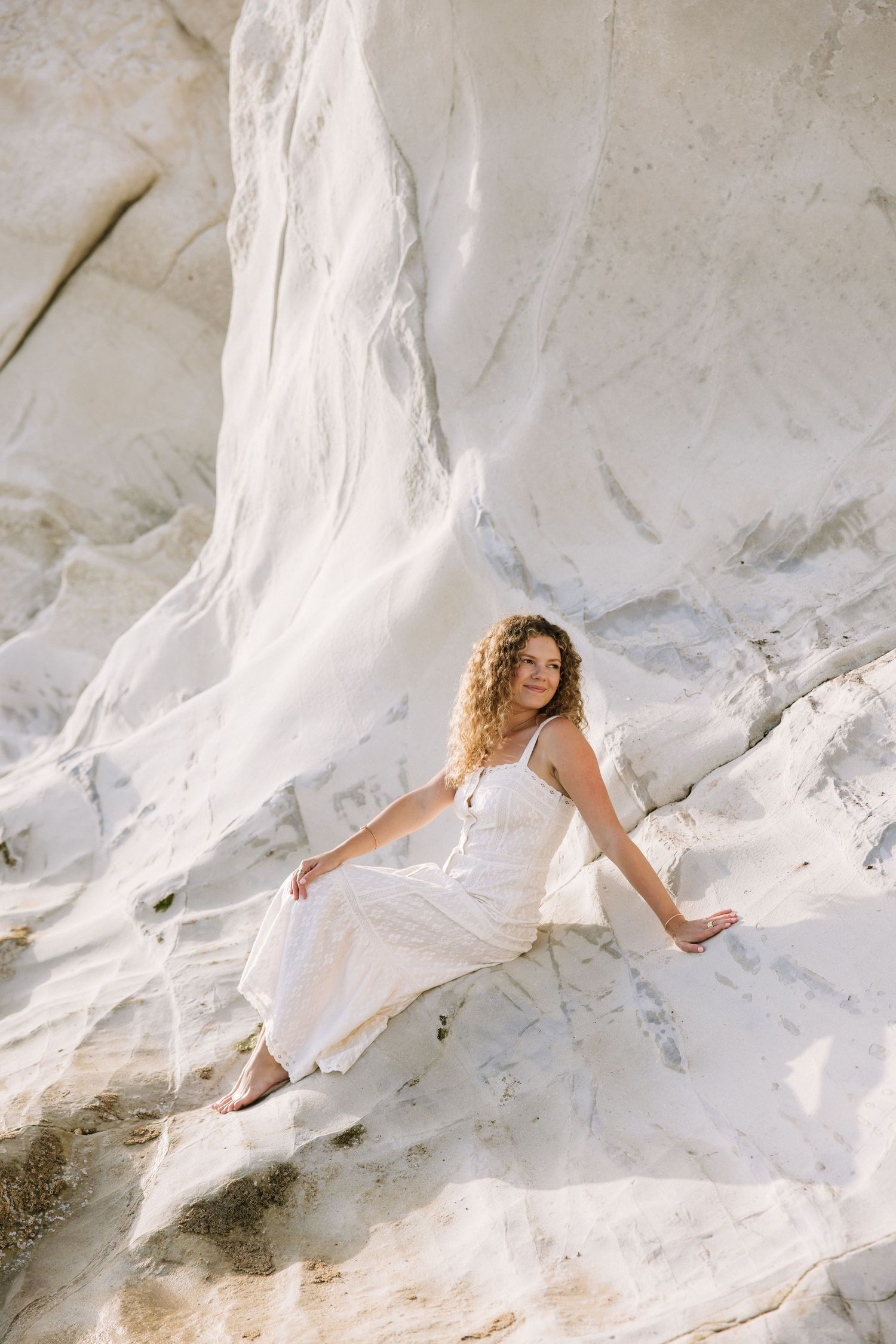 Woman in white dress sits gracefully against white rock cliffs in natural sunlight.
