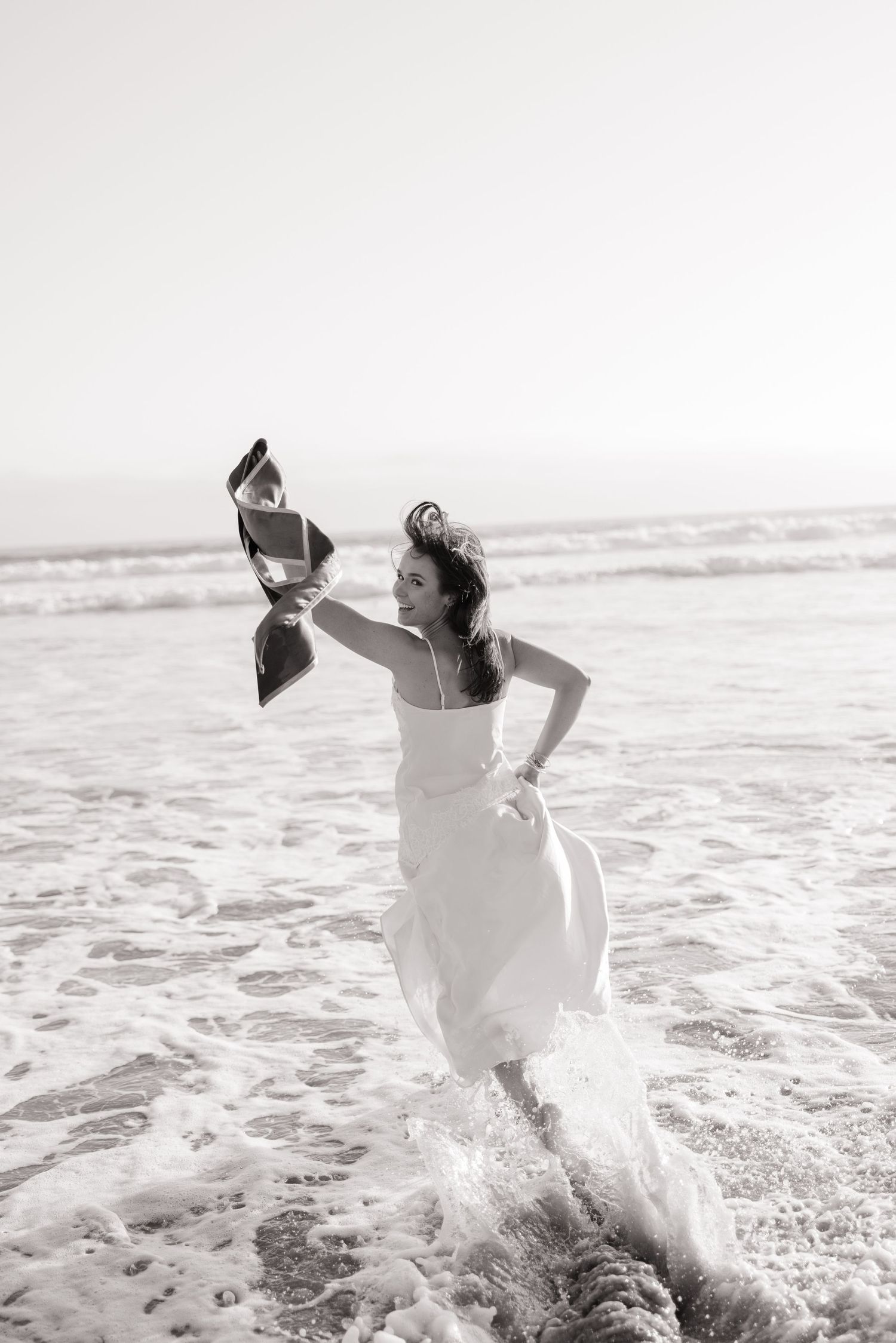 Black and white photo of woman in white dress running through ocean waves with graduation cap.
