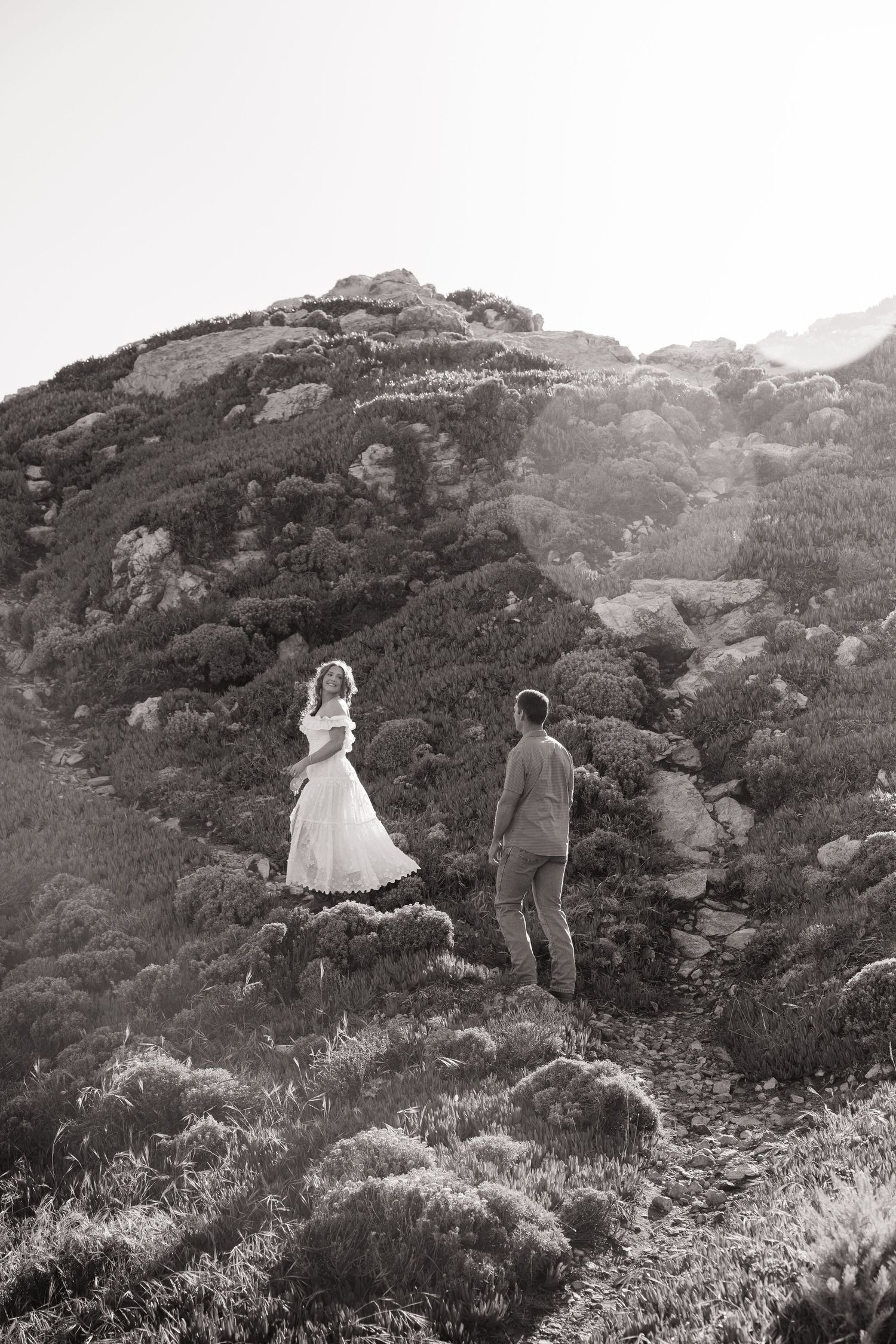 Black and white photo of a couple standing on a rugged hillside trail surrounded by wild shrubs.