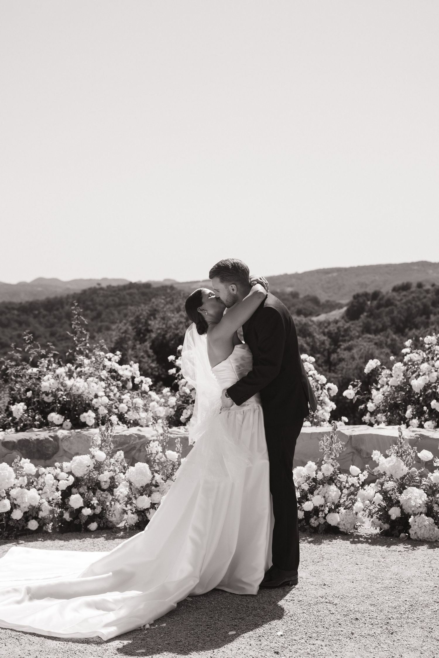 Bride and groom share first kiss at outdoor wedding ceremony surrounded by white floral arrangements.