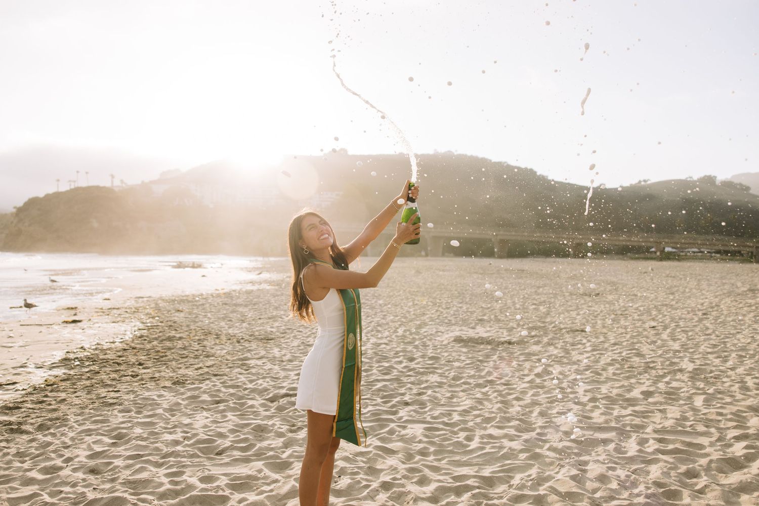 Joyful woman in white dress pops champagne bottle on sunny beach with mountains in background.